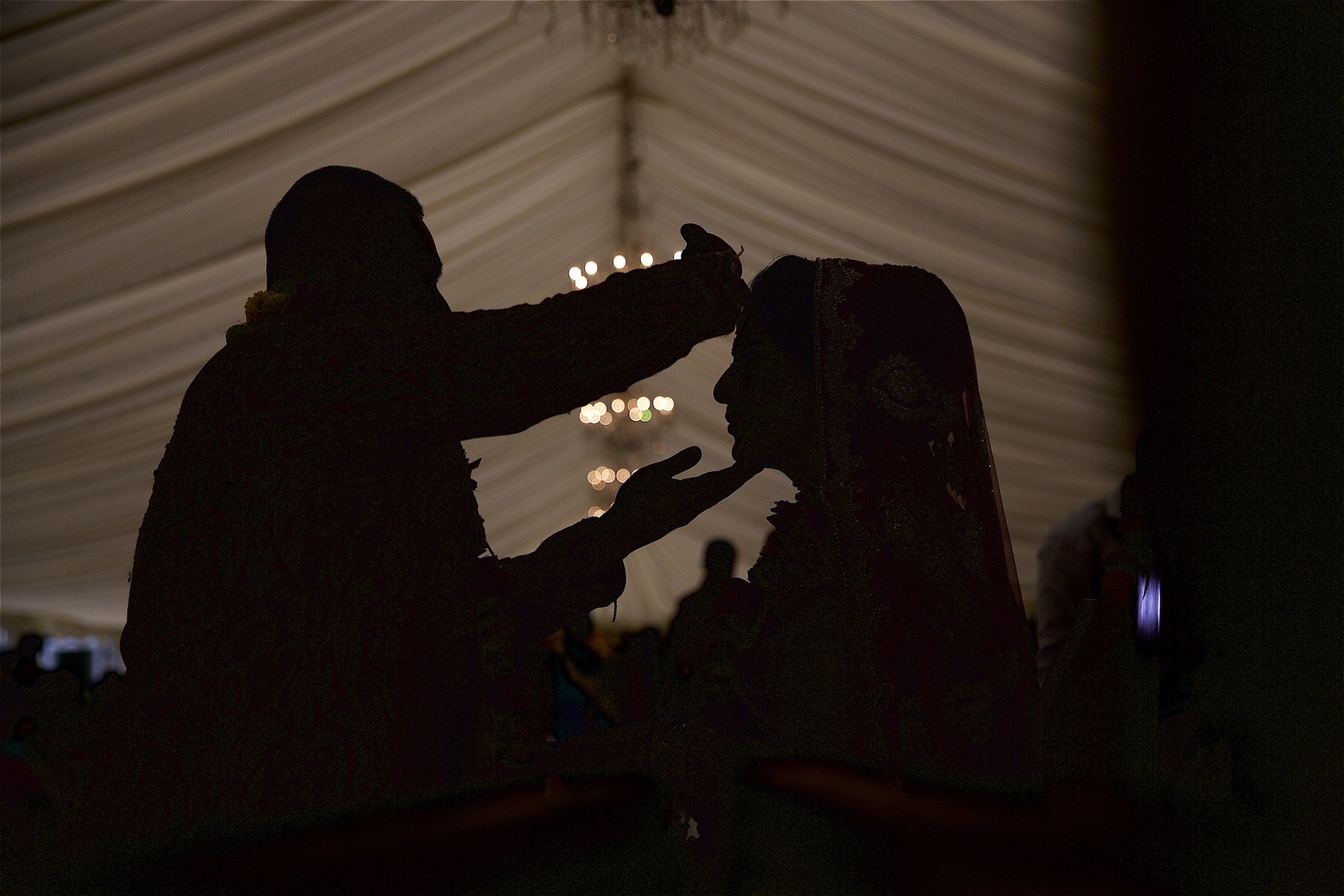 Silhouette of a man placing a tiara on a woman's head at a wedding reception inside a tent with draped ceiling and hanging lights.