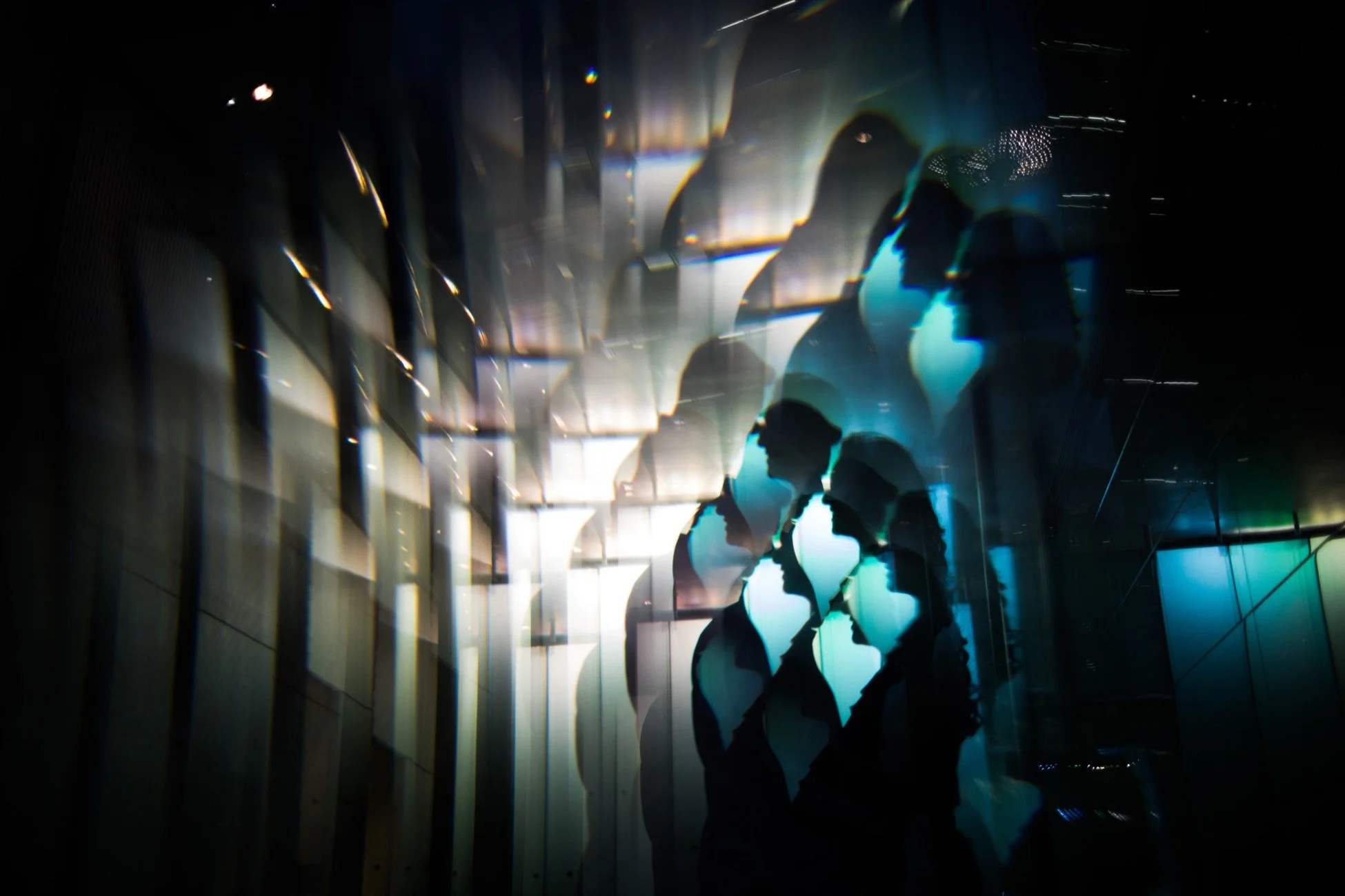 Silhouettes of five people standing in a dark room with bright, colorful lights reflecting off a wall. The scene has an artistic, abstract quality with a focus on shadows and reflections.