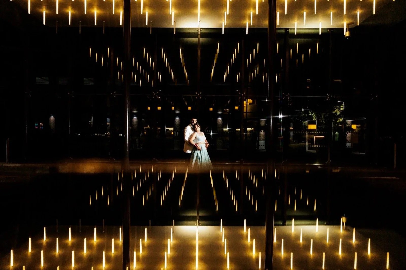 A couple standing together in front of a large glass wall with glowing vertical lights, reflections on the shiny floor, and a dark background.