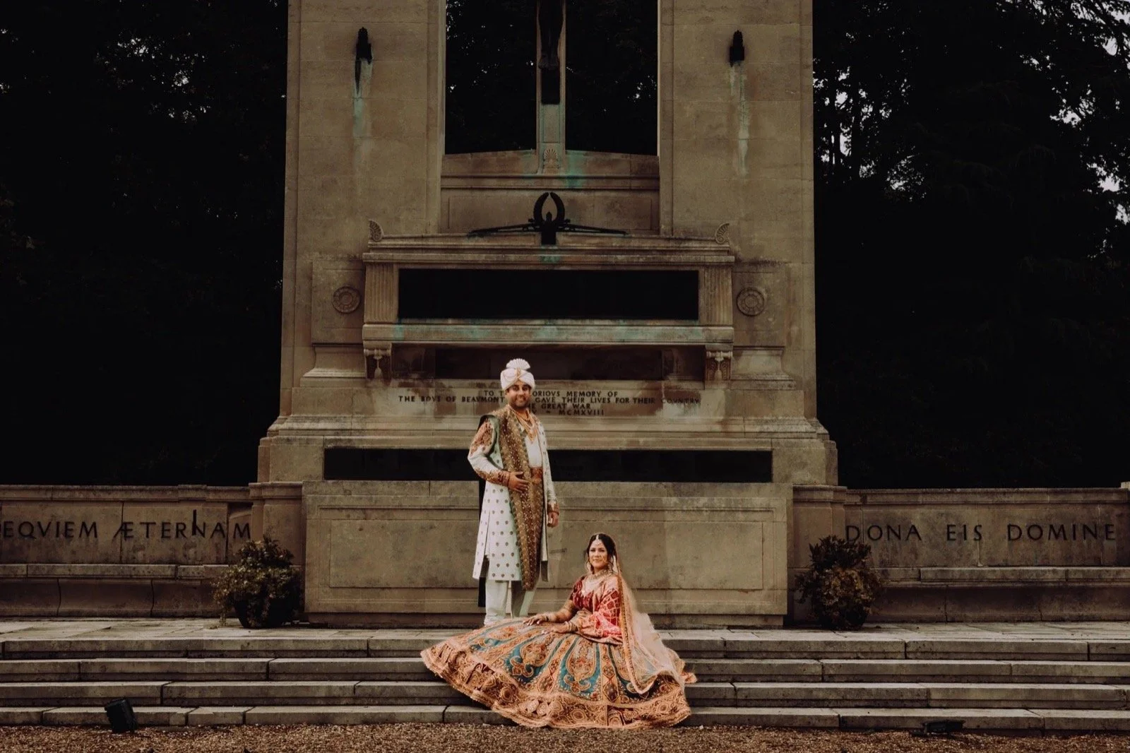 A man and woman in traditional Indian attire pose in front of a large stone monument with steps, inscriptions, and a clock, during dusk or evening.