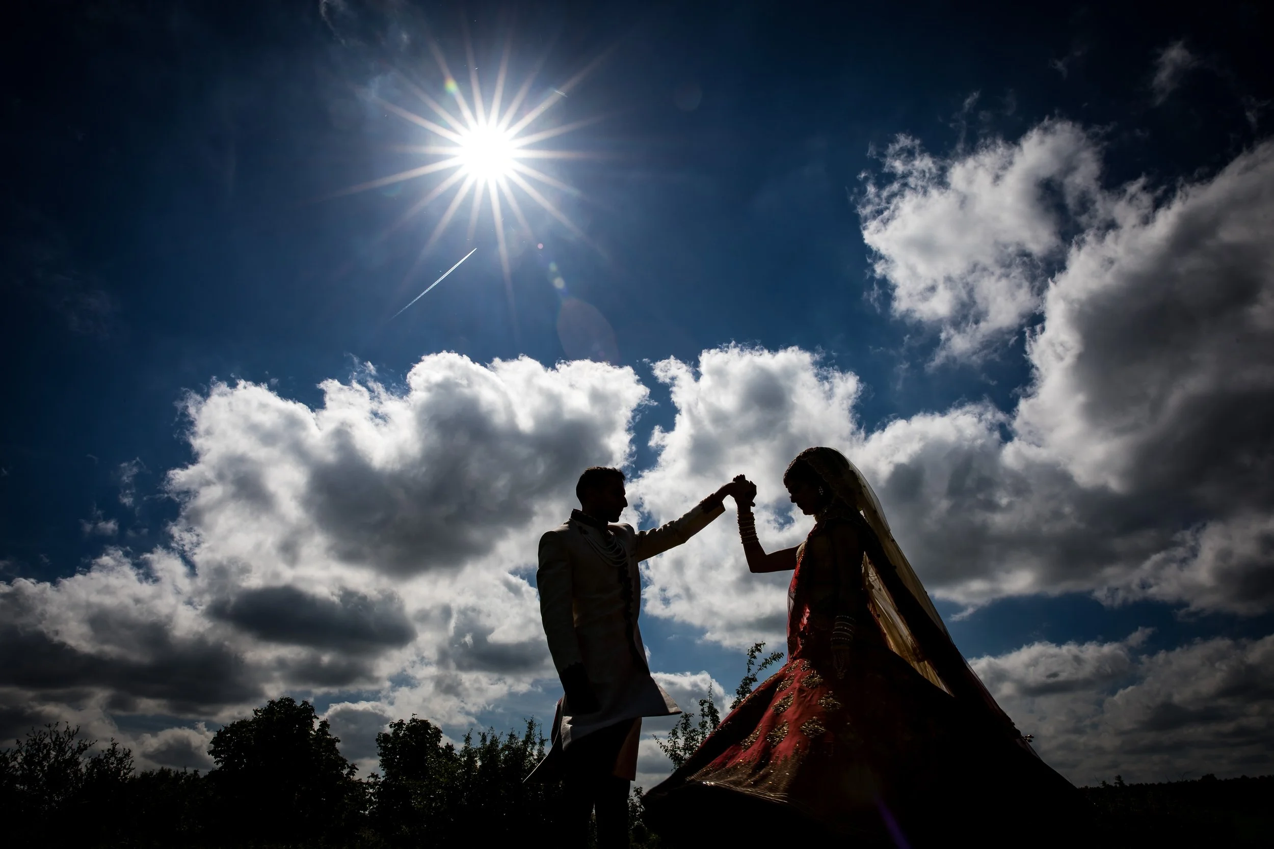 Silhouette of a couple, with a man lifting a woman’s hand, against a bright sun and cloudy sky, appearing to be outdoors during daytime.