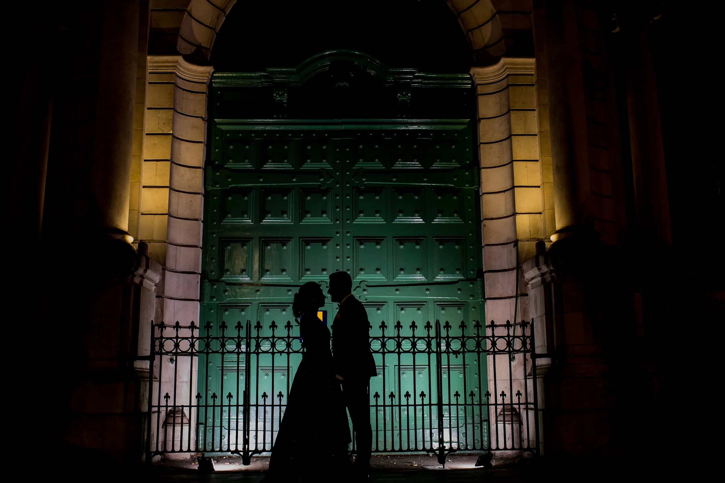 Silhouettes of a man and woman standing close together in front of a large green door with an arched top, surrounded by stone columns and a decorative fence, in low lighting.