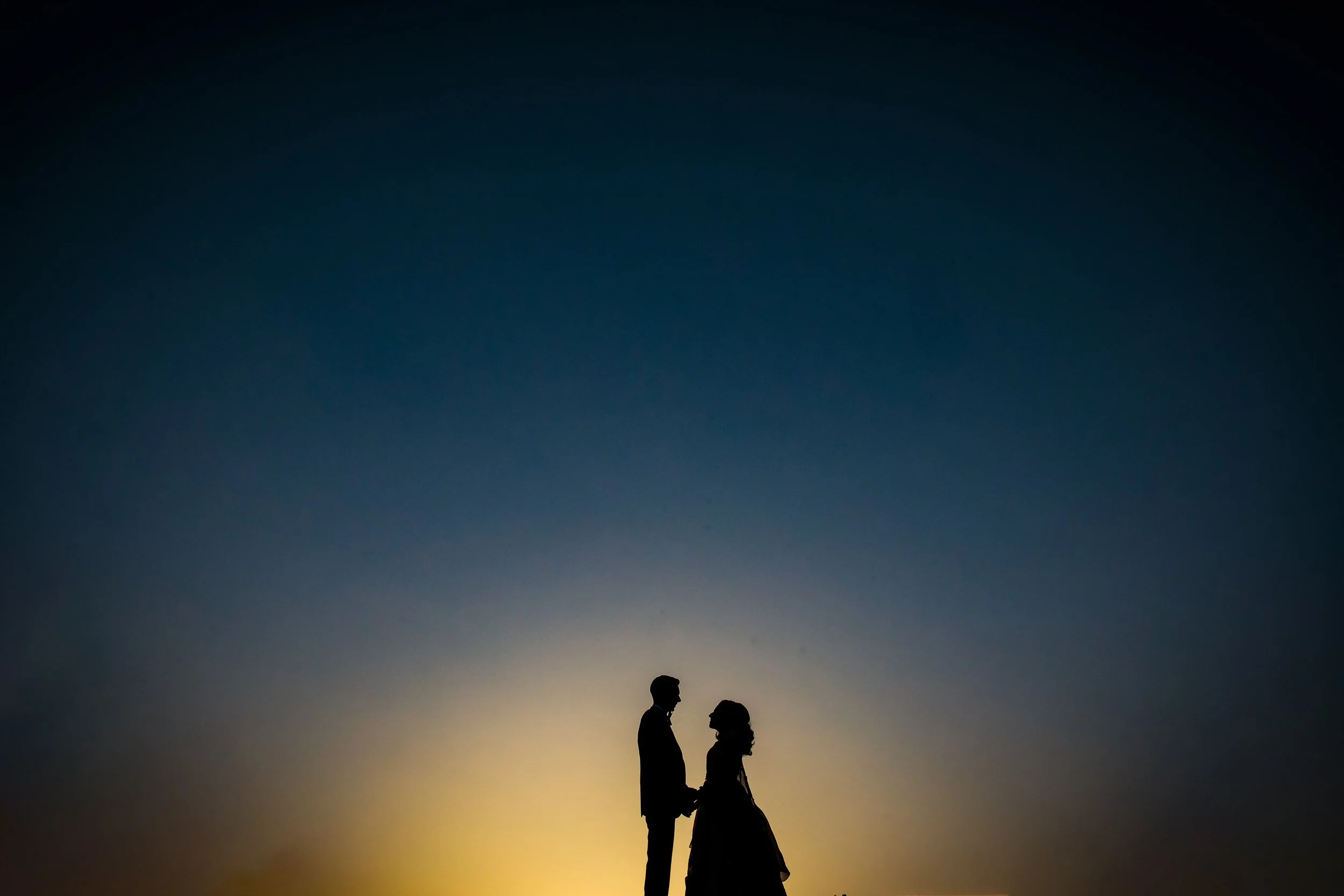 Silhouette of a couple holding hands and facing each other at sunset or sunrise, with a gradient sky in shades of dark blue and yellow.