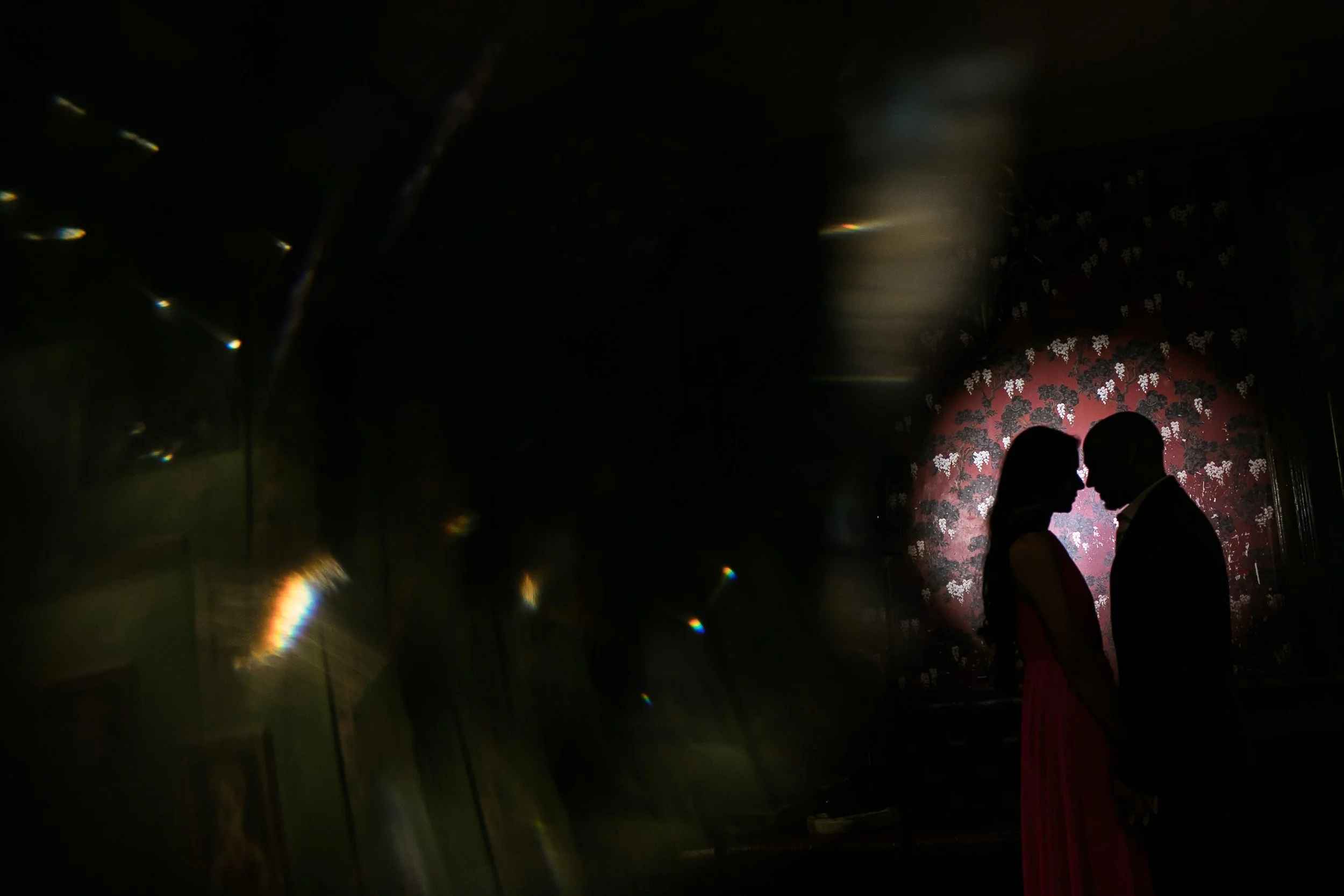 Silhouette of a couple kissing in front of a red floral-patterned wall in a dimly lit room.
