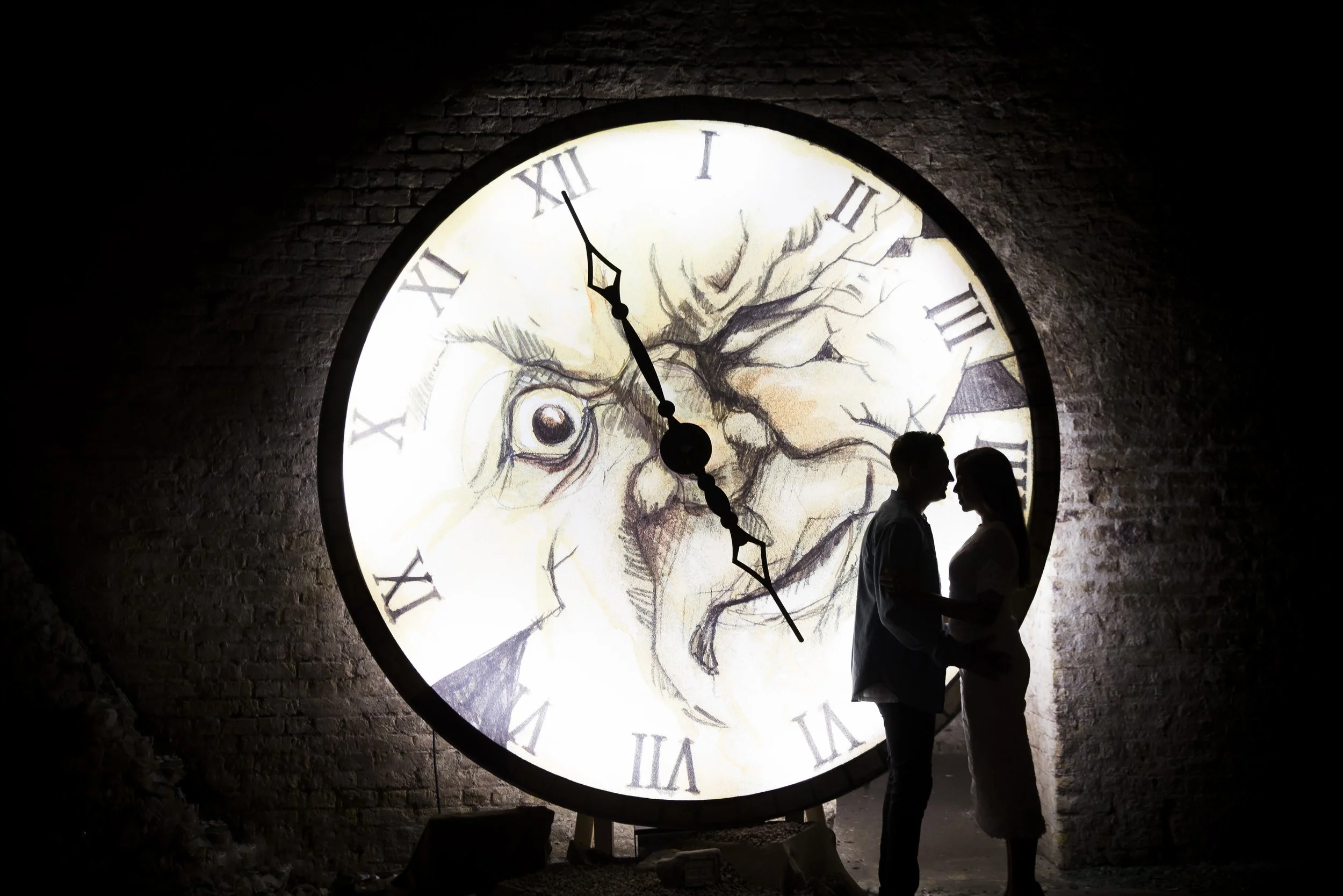 A large illuminated clock with Roman numerals on a brick wall, featuring a detailed artistic face of a humanoid squirrel, with silhouettes of a man and a woman standing closely and looking at each other in front of the clock at night.
