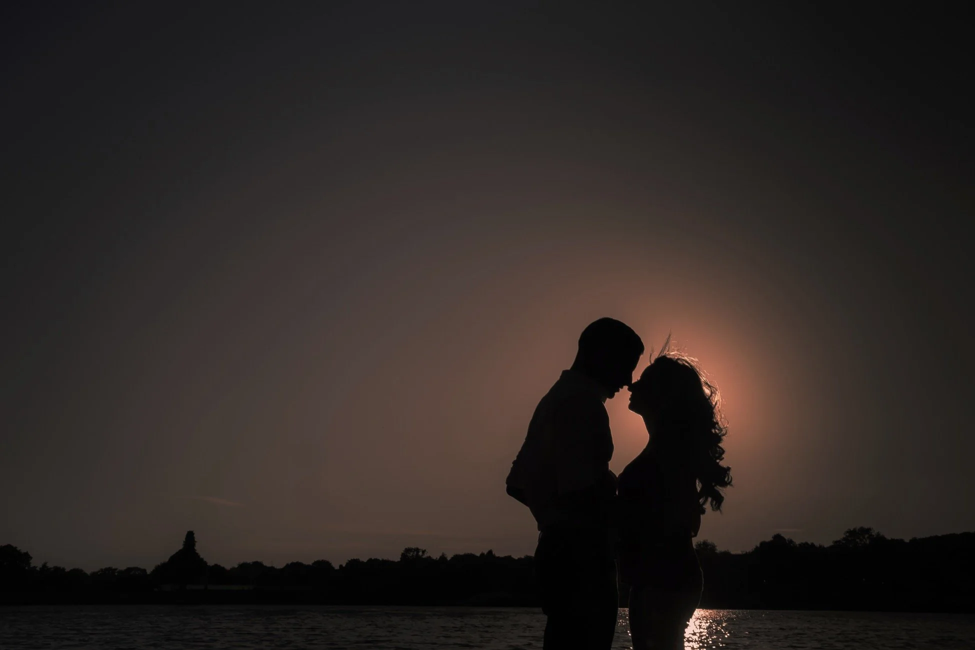 Silhouette of a couple embracing and touching foreheads against a sunset background over water, with trees and a distant pagoda in the landscape.
