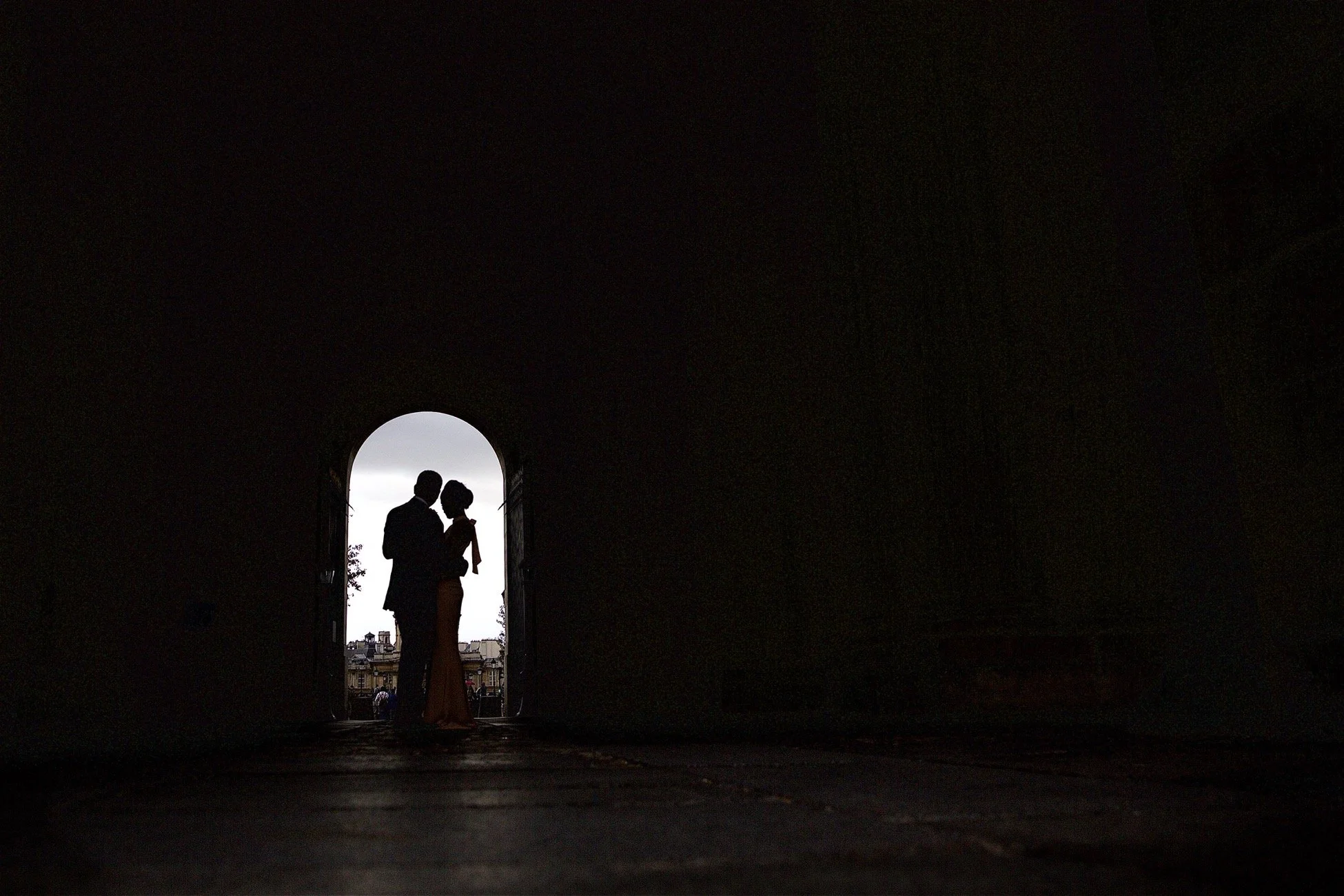 Silhouette of a couple standing close together in an arched doorway, with a cityscape view in the background, during dusk or night.