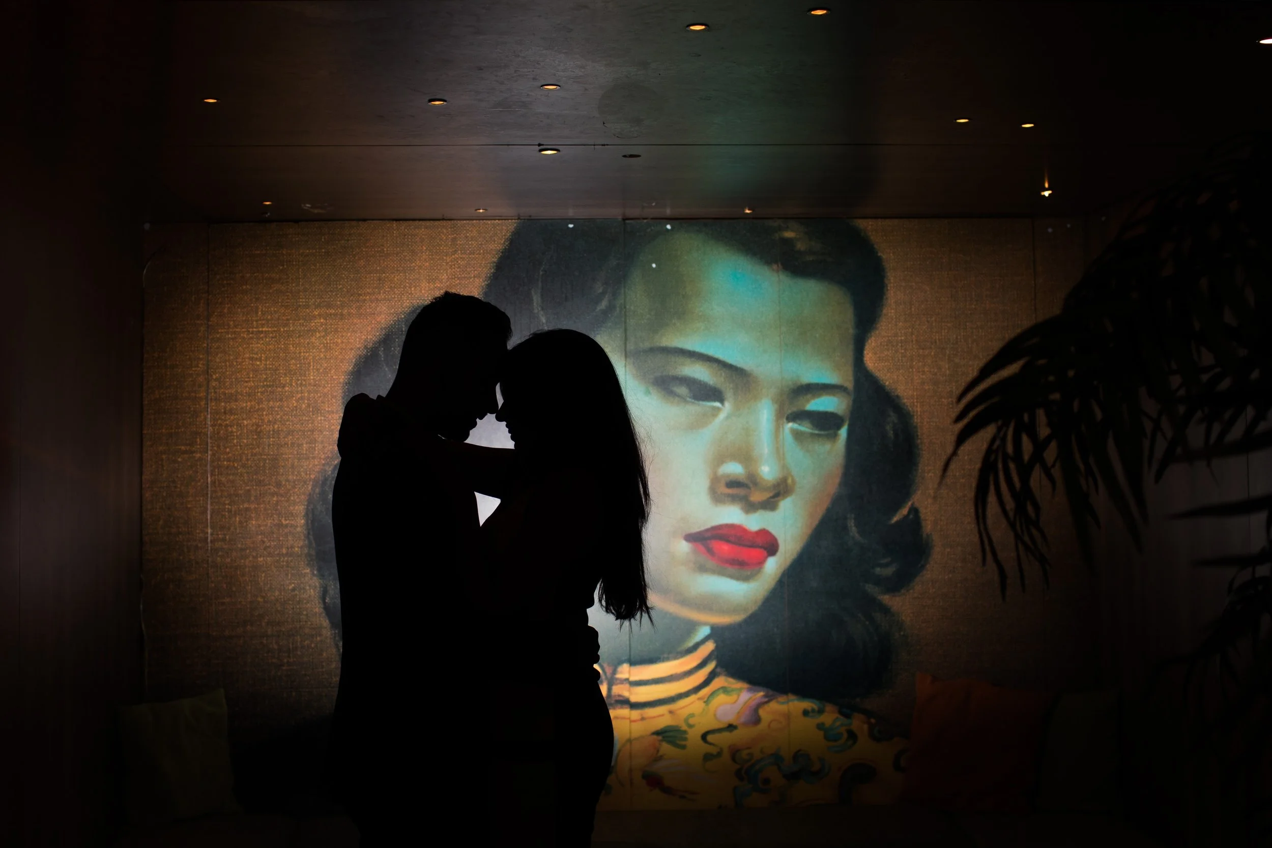 Silhouetted couple kissing in front of a large portrait of a woman with dark hair, red lips, and a striped shirt, projected on a wall in a dimly lit room.