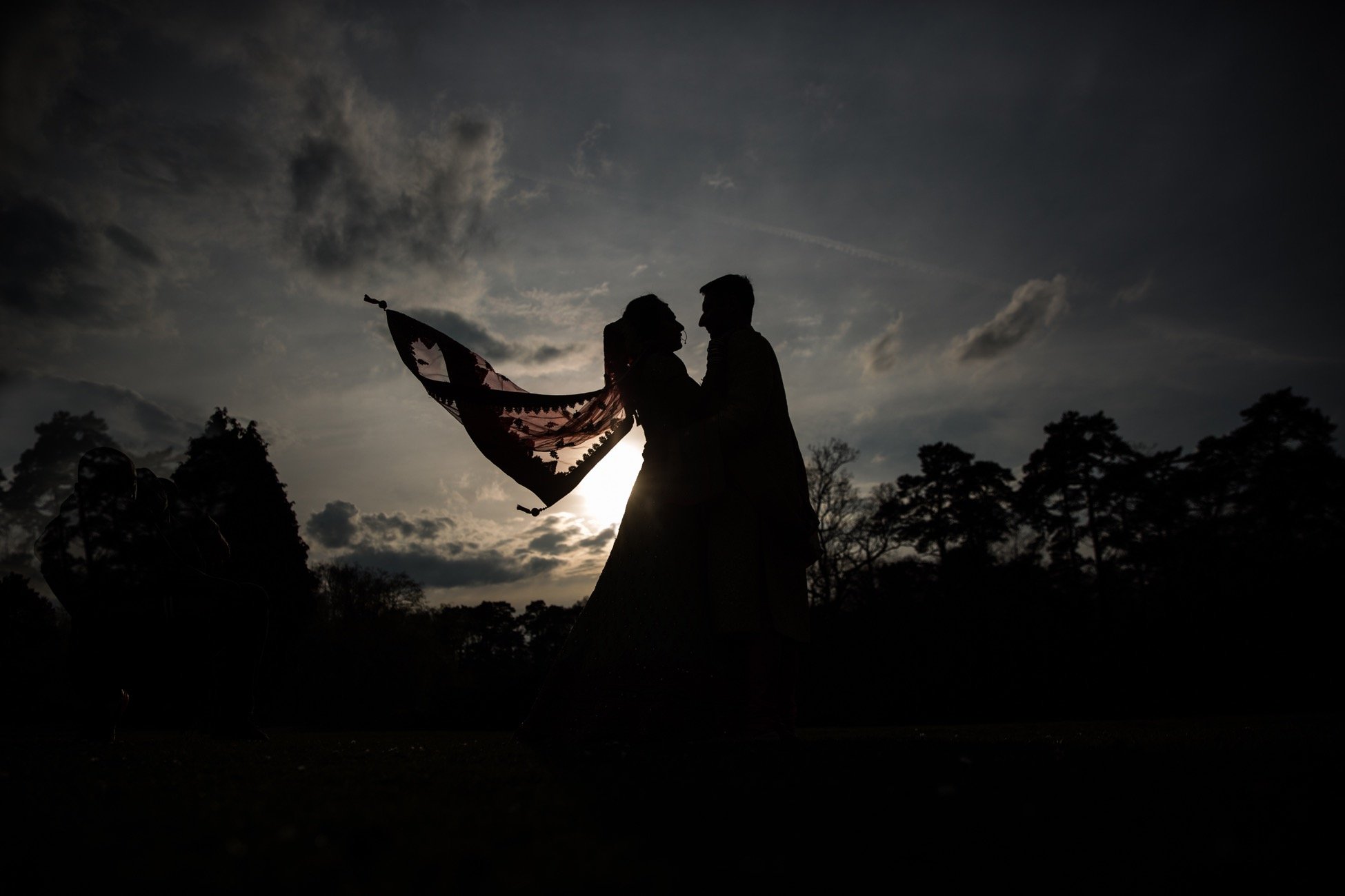 Silhouette of a couple holding a flag under a cloudy sky at sunset, with trees in the background.