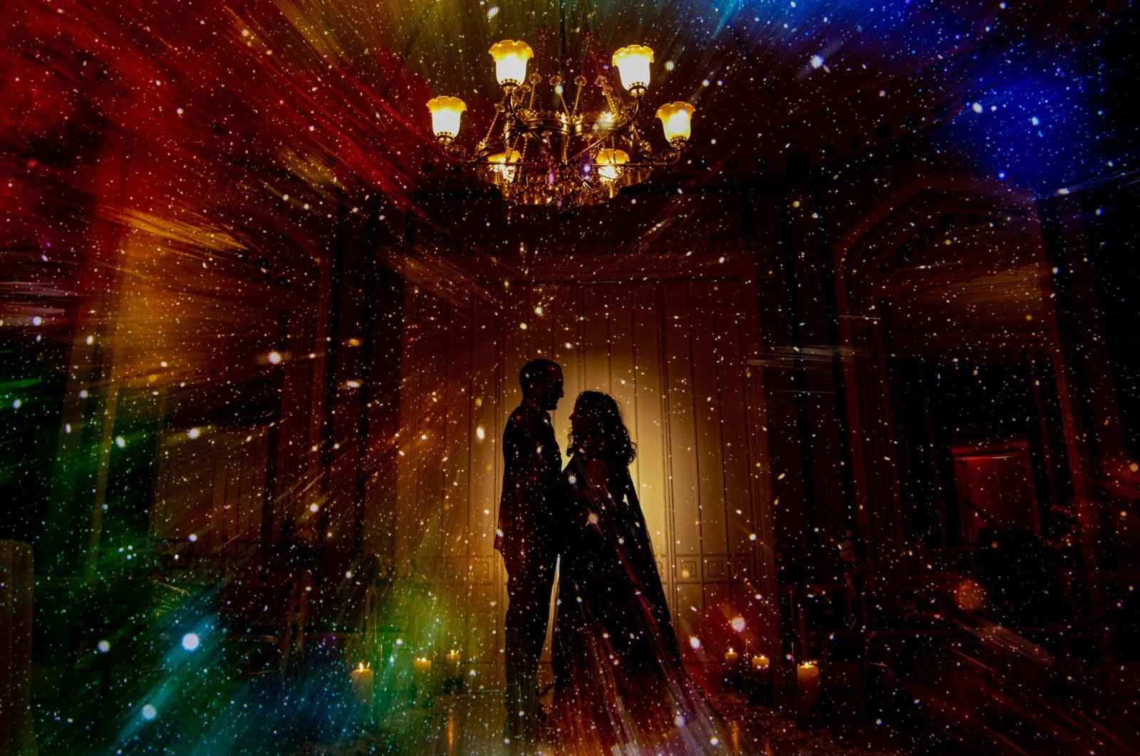 Silhouettes of a man and woman standing close together in a dimly lit room with a chandelier on the ceiling, surrounded by colorful, star-like specks of light.