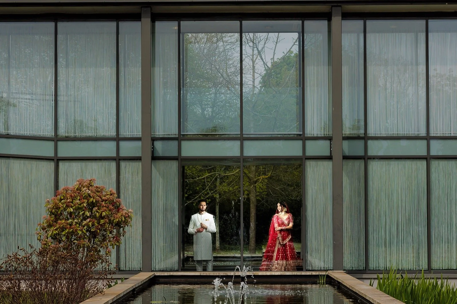 A man and woman dressed in traditional Indian attire are inside a modern glass building, standing near a large window with trees visible outside. The woman is wearing a red and gold saree, and the man is in a white sherwani. They are positioned behind a water fountain with plants and flowers in the foreground.