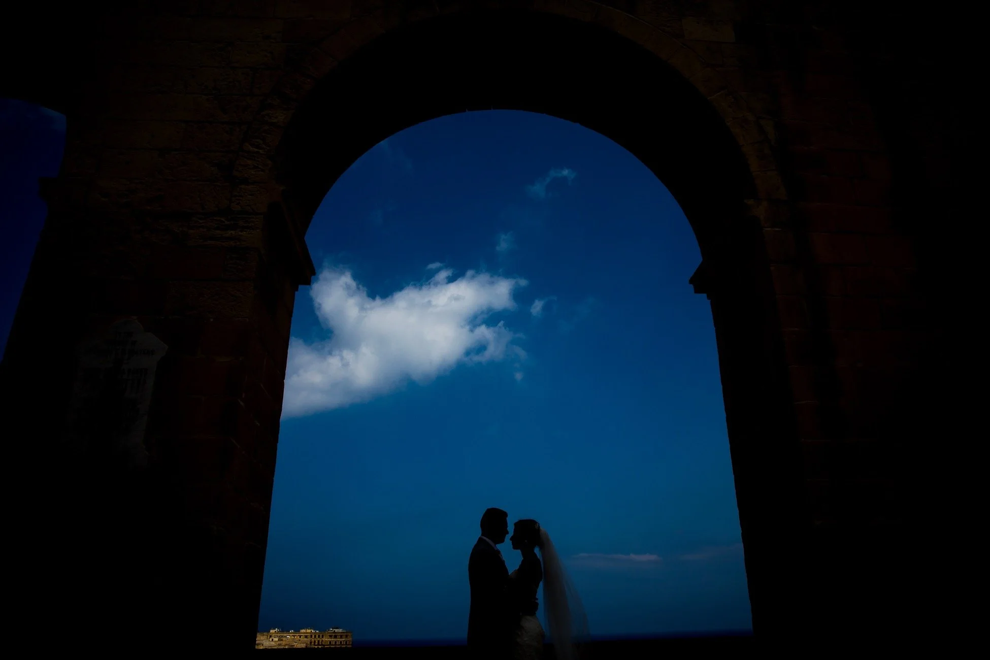 Silhouette of a newlywed couple kissing beneath an archway with a blue sky and clouds in the background.