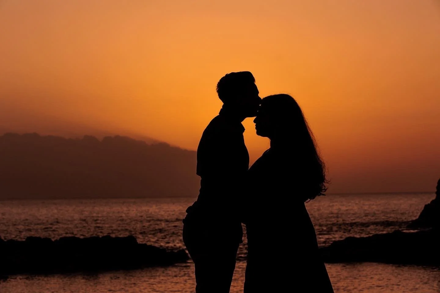 Silhouettes of a couple standing close and touching noses at sunset by the water, with a colorful sky and rocky shoreline in the background.