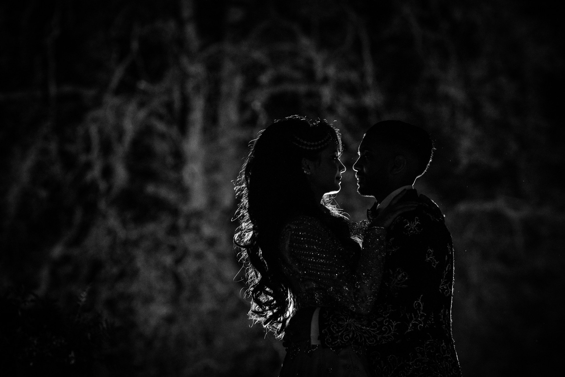 A black and white photo of a couple standing close, facing each other, with their faces almost touching. The background appears dark and textured.