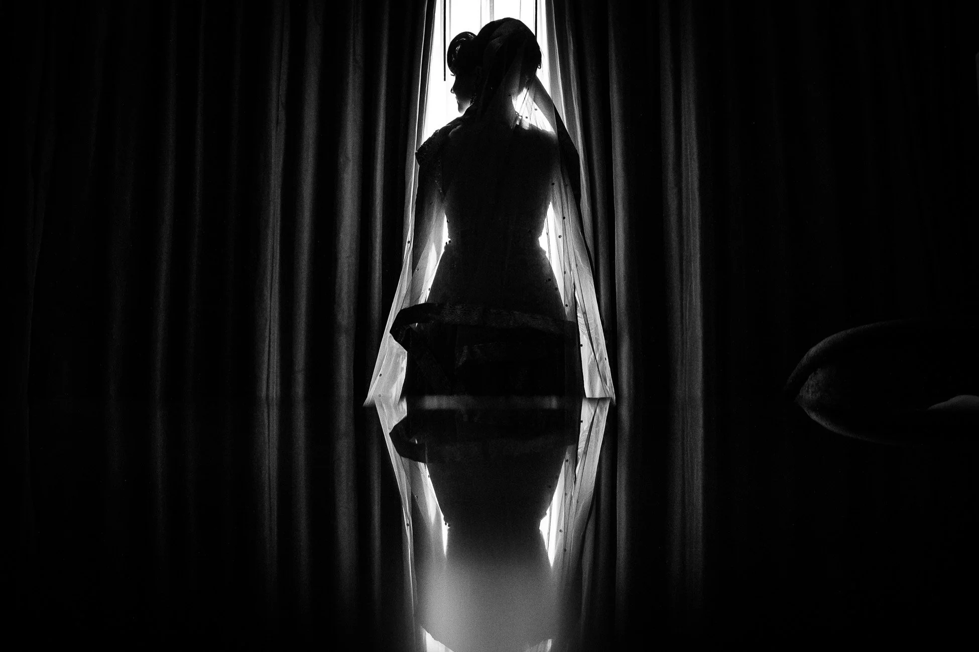 Silhouette of a woman in a wedding dress and veil, standing against a window with curtains, casting a reflection on a shiny surface below.