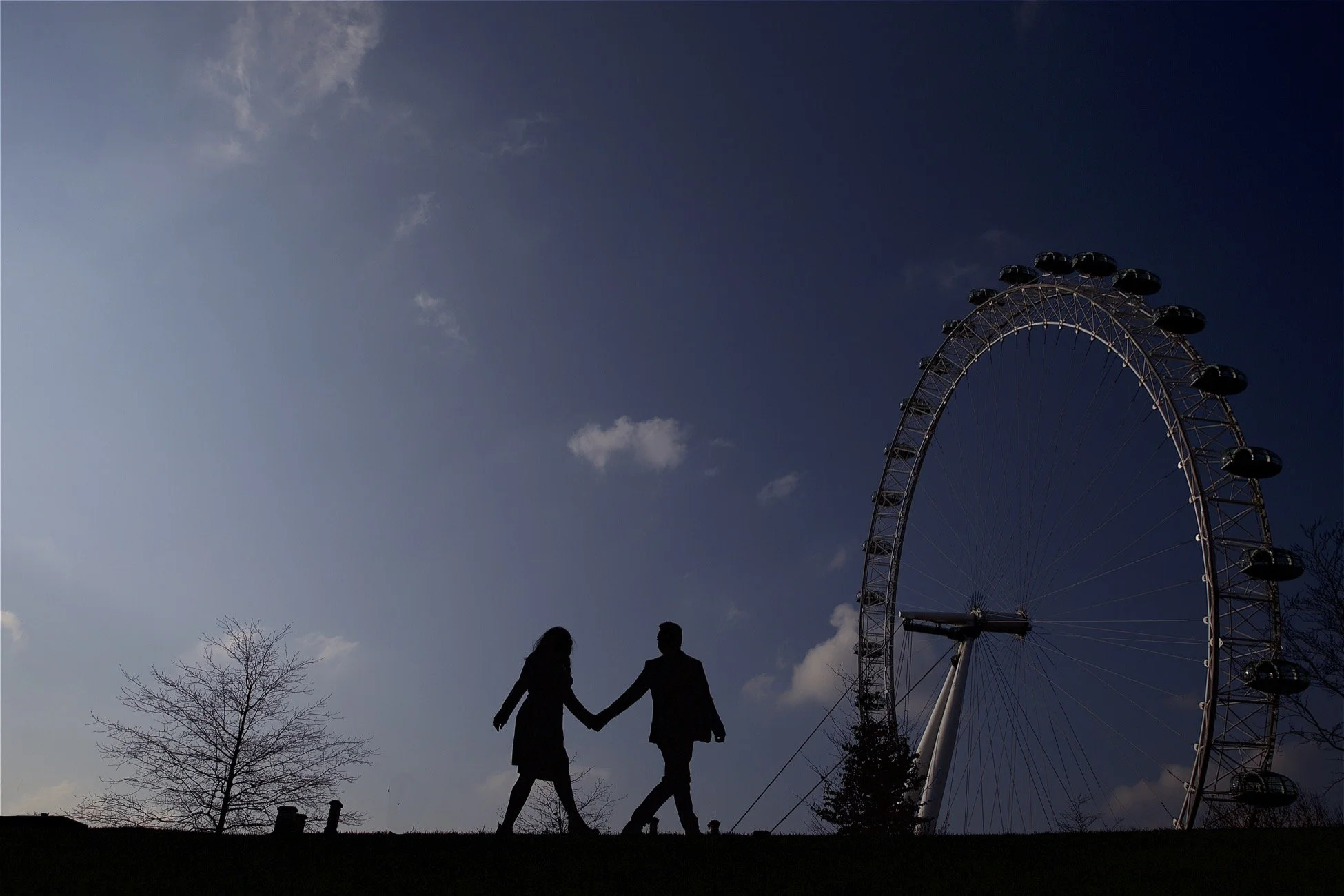 Silhouettes of a man and woman holding hands, standing on a hill with a large Ferris wheel in the background against a partly cloudy sky.