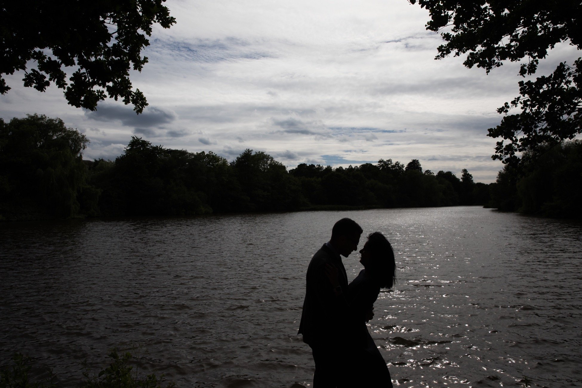 Silhouettes of a couple holding hands by a river with trees and a cloudy sky in the background.