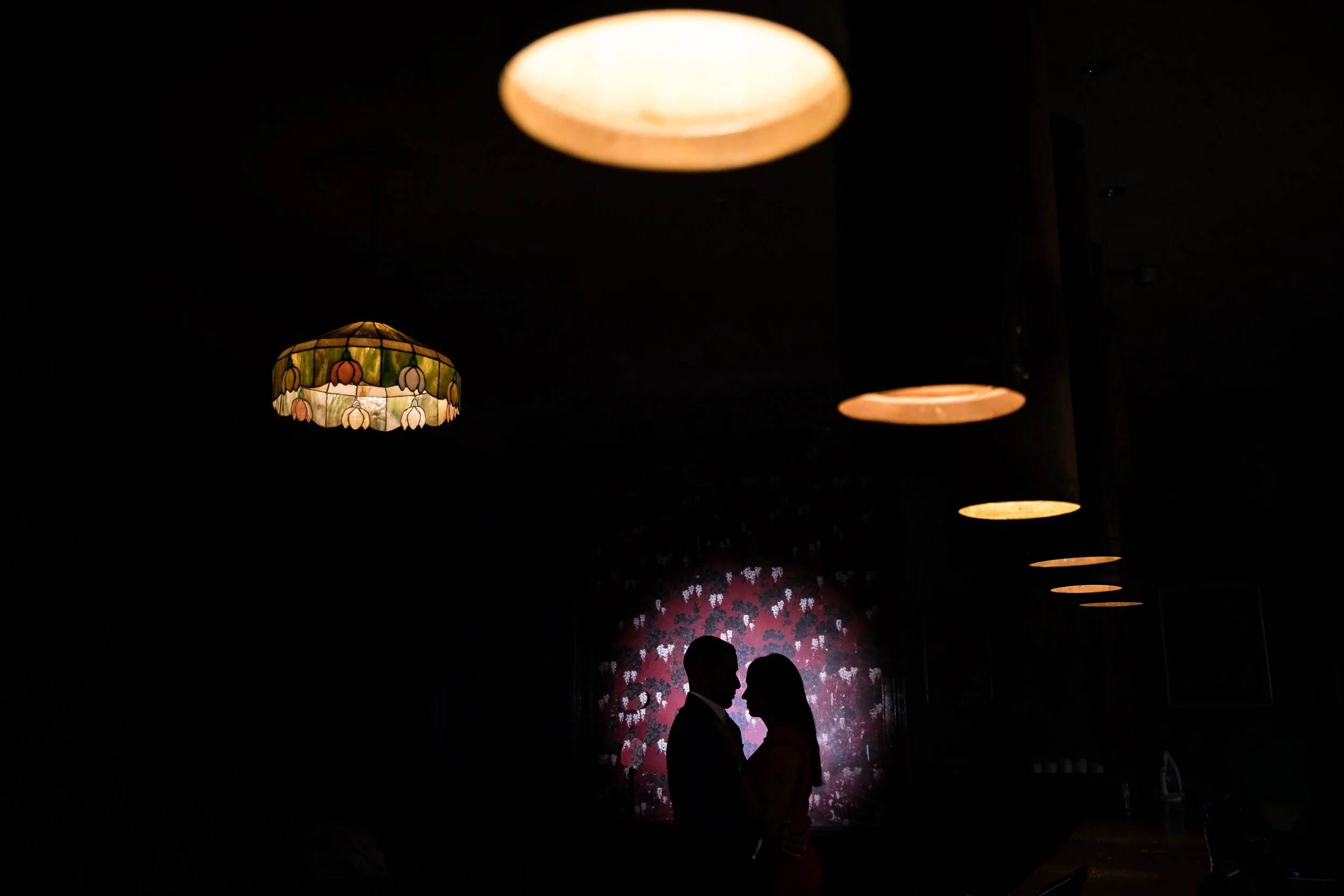 Dimly lit interior with hanging ceiling lights and two people silhouetted against a floral-patterned wall.