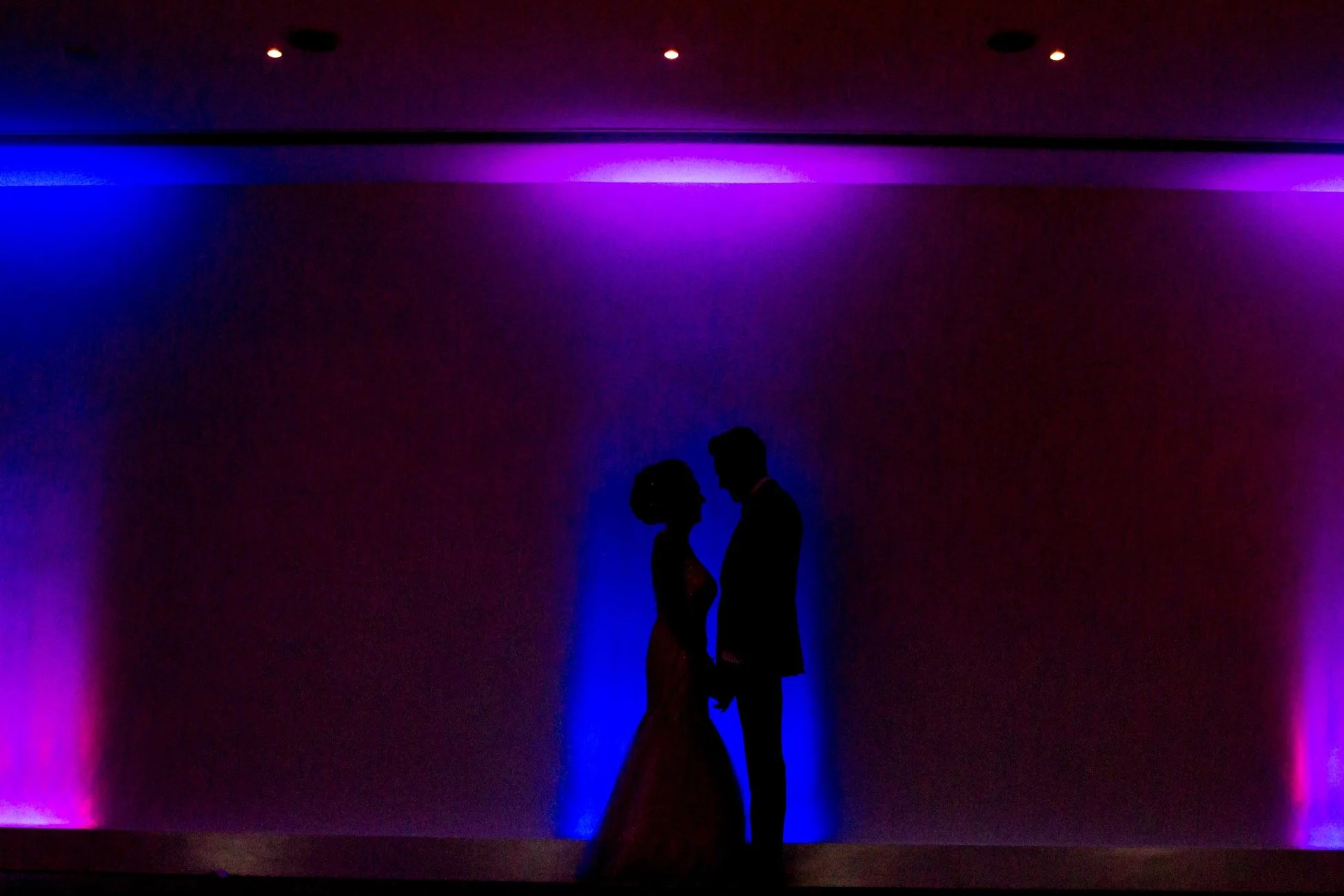 Silhouettes of a man and woman close together in front of a colorful purple and blue lit background.