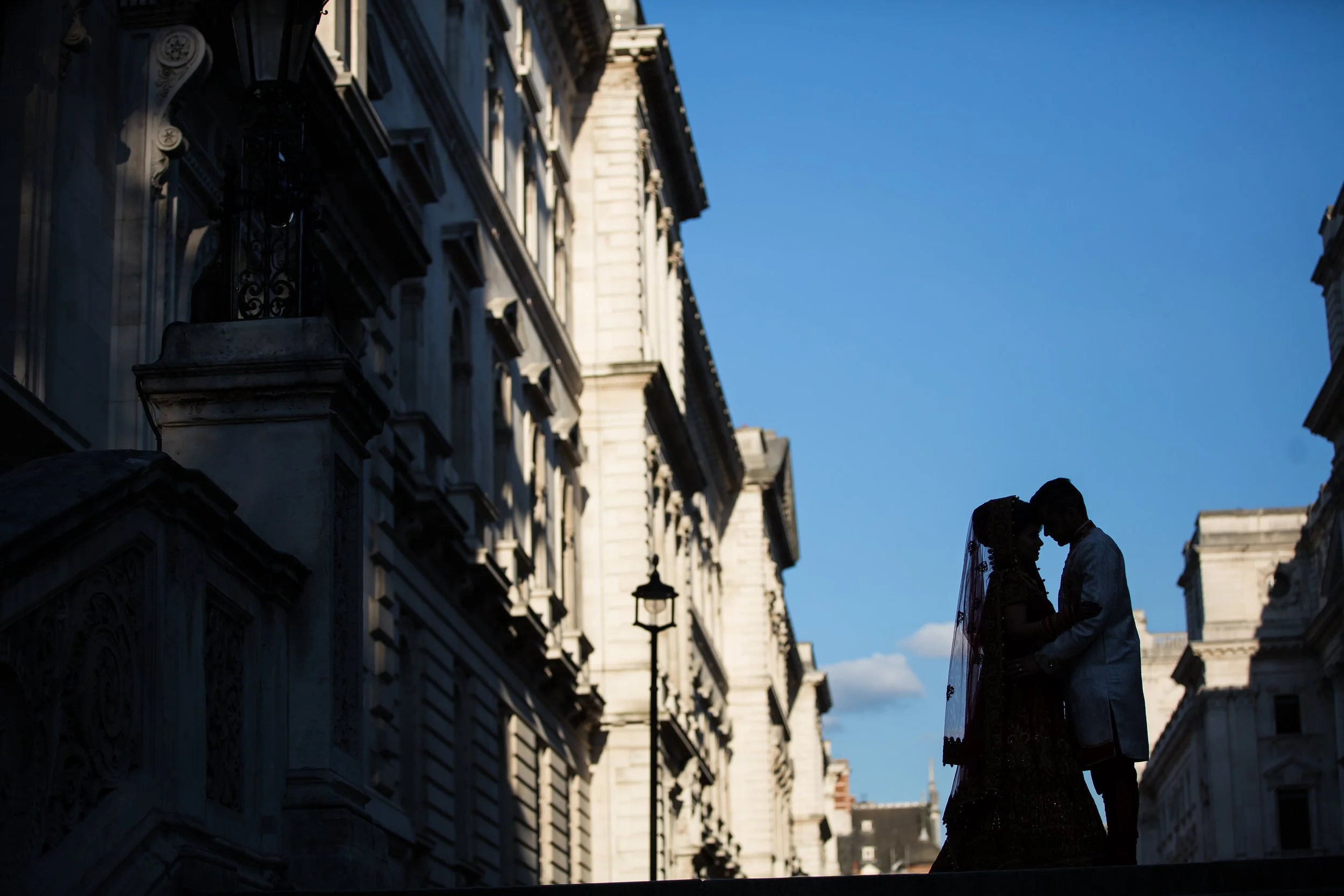 A silhouette of a couple, with a woman in a wedding dress and a man in a suit, standing close together on a building ledge, facing each other against a backdrop of historic Paris buildings and a blue sky.