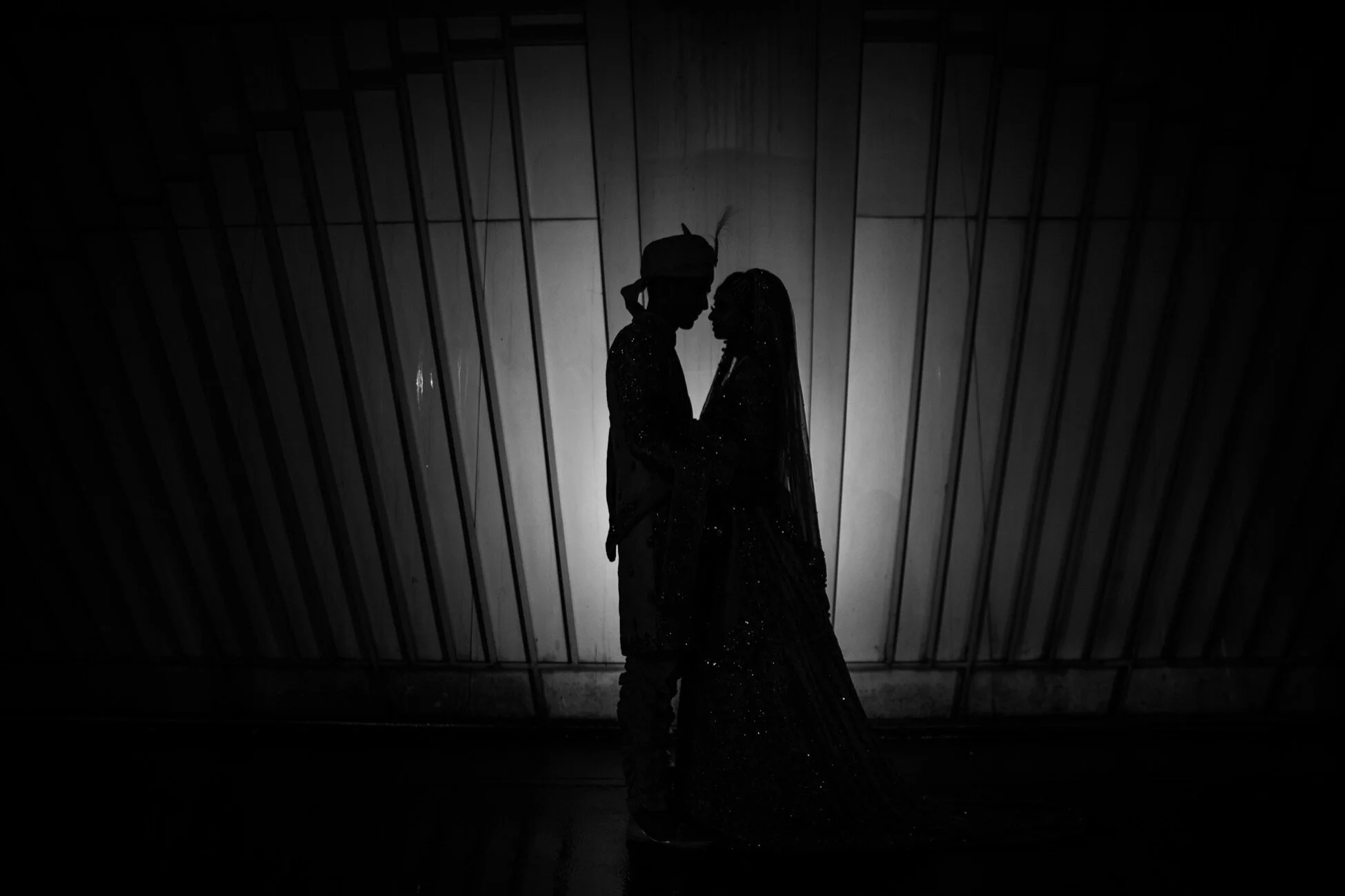 Silhouette of a couple in traditional Indian wedding attire facing each other, standing closely in front of a wooden wall.
