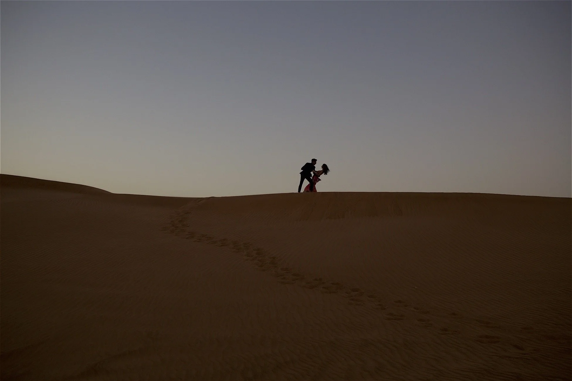 Silhouettes of a man and a woman holding hands on a sandy desert dune at sunset or twilight.