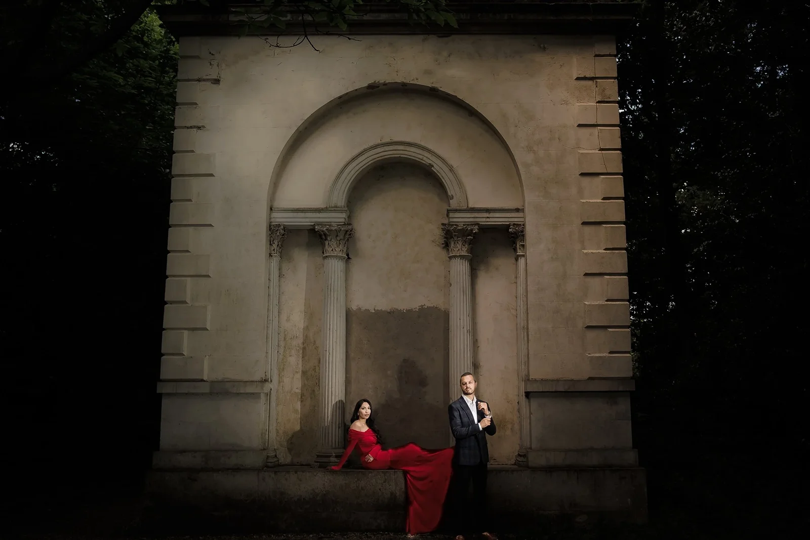 A man in a dark suit holding a drink, and a woman in a red dress sitting against a classical architectural structure with columns, within a dark outdoor setting.