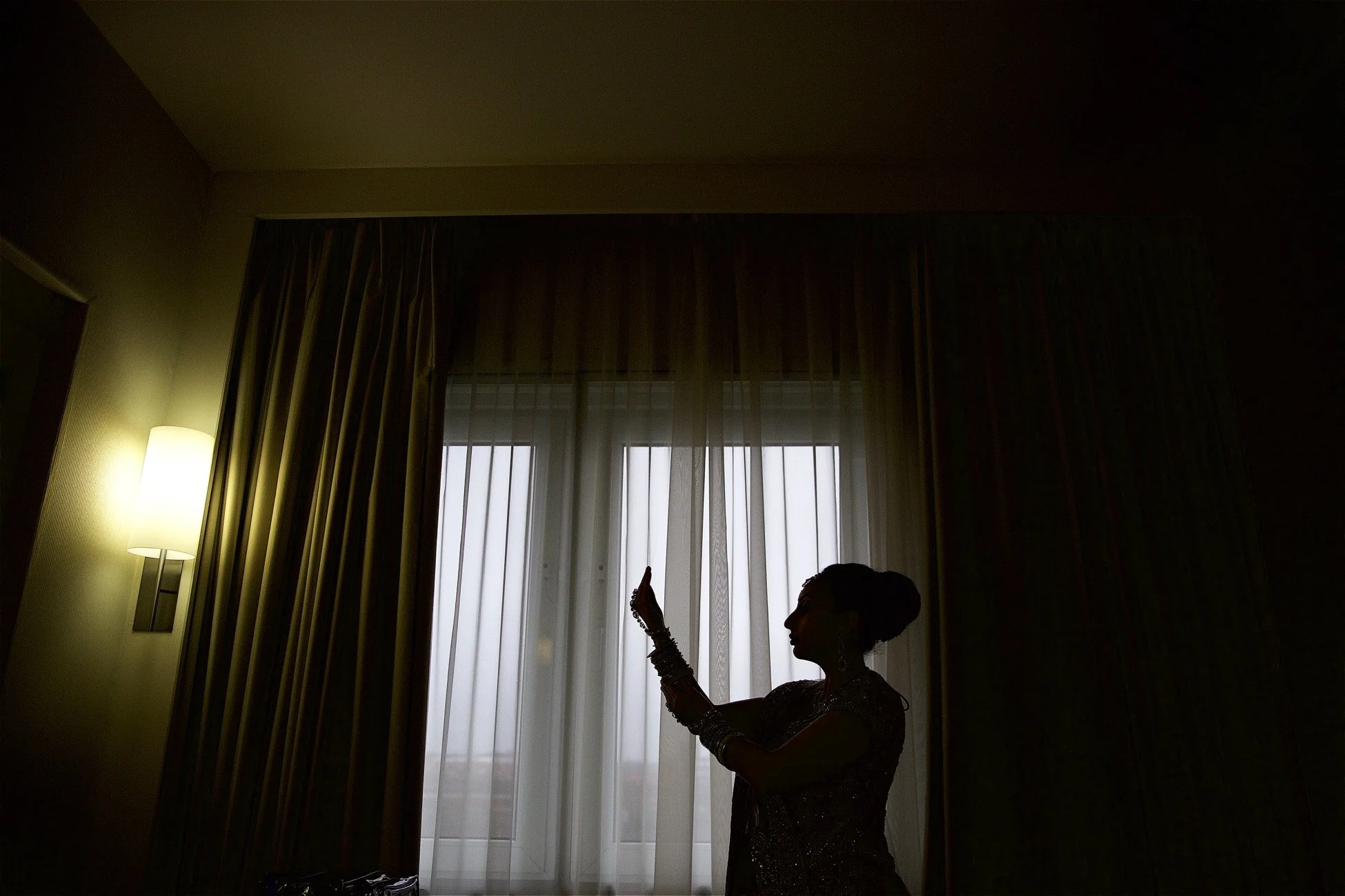 Silhouette of a woman inside a room with a large window and curtains, a standing lamp on the left, with light streaming through the curtains.