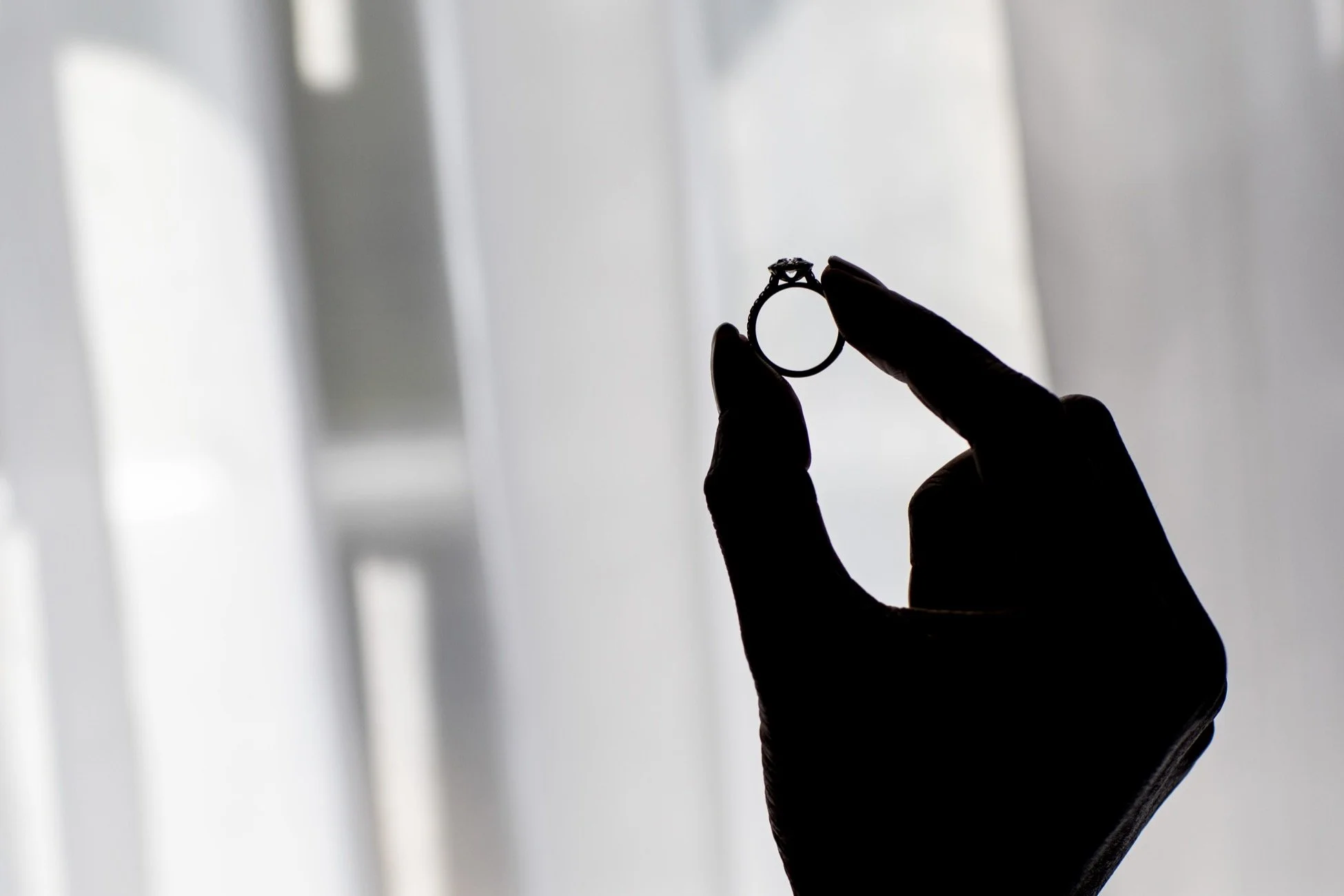 Silhouette of a hand with black glove holding an engagement ring against a softly lit background.
