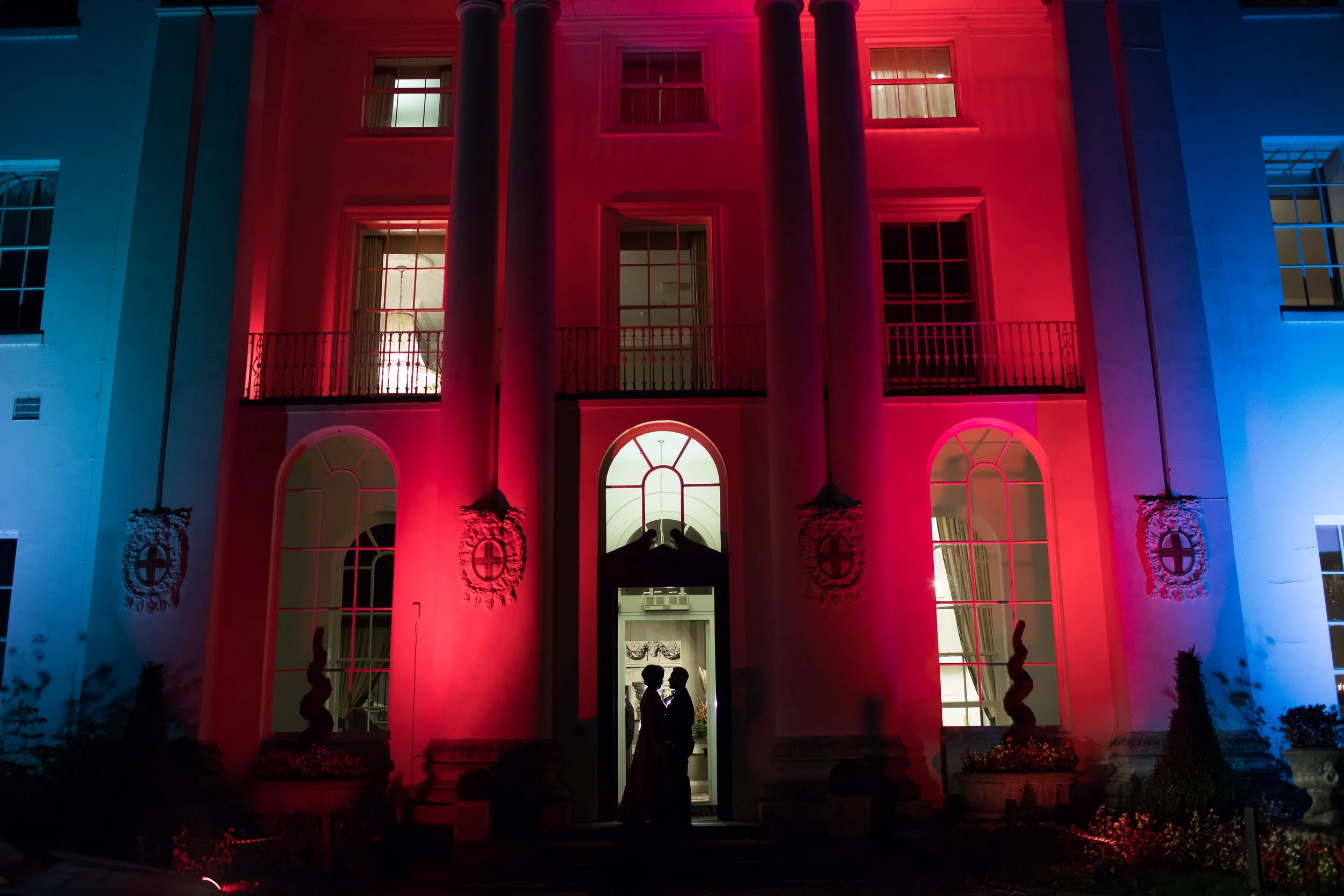 The front of a large, elegant house illuminated with red, white, and blue lights at night. The house has tall columns, arched windows, and a staircase leading to the door. Two silhouetted figures are visible at the entrance.