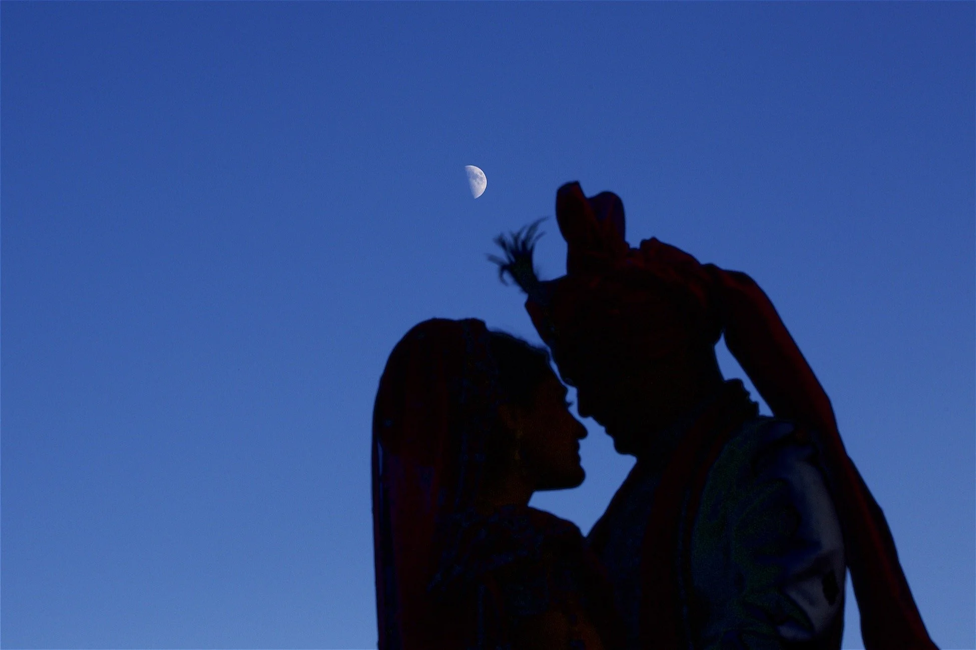 Silhouette of two people close together, touching foreheads, with a blue sky and moon in the background.