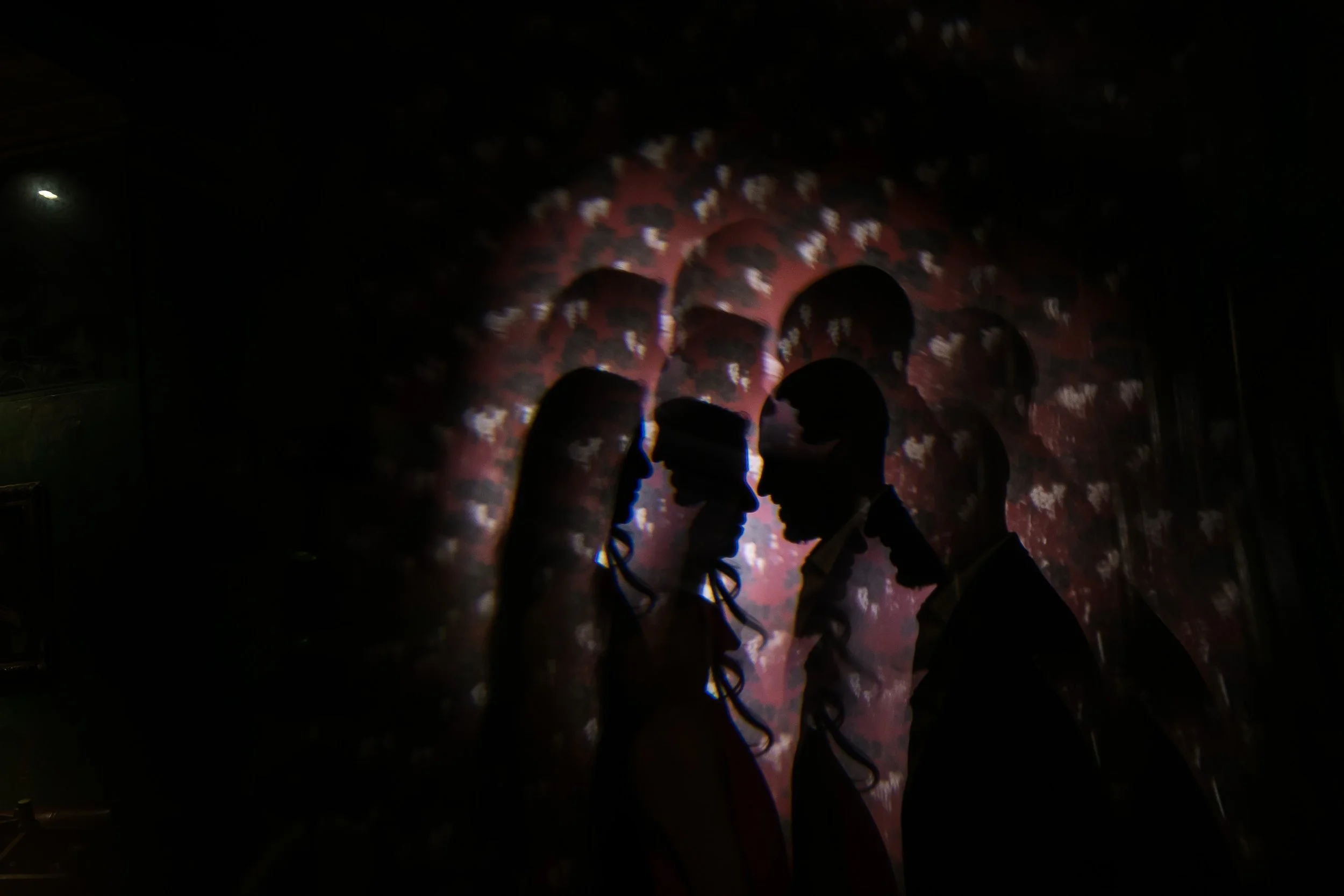 Silhouettes of three people facing each other in front of a red patterned backdrop with light reflections, possibly at night or in a dimly lit room.