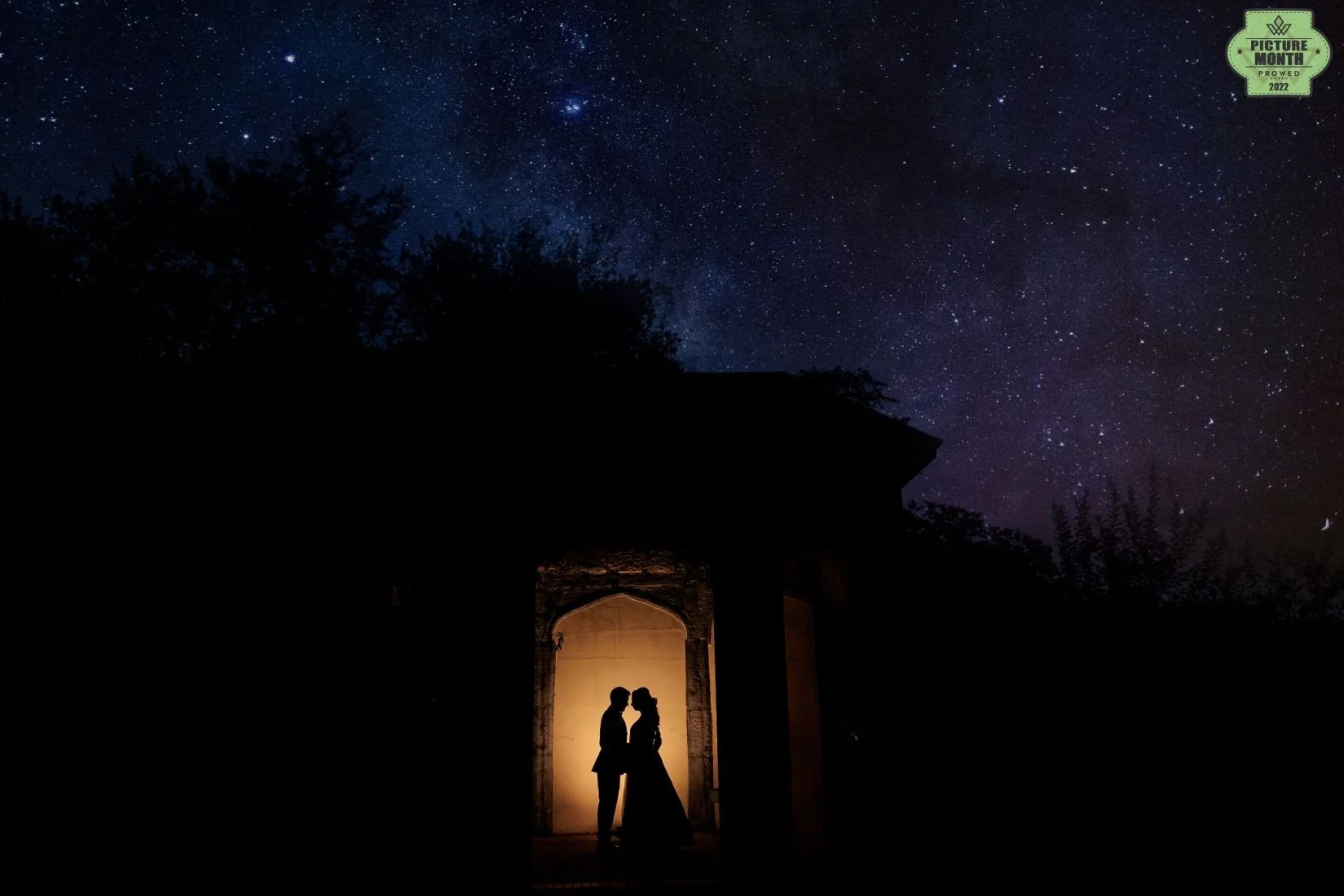 Silhouette of a couple standing close together under an archway, illuminated from within, against a backdrop of a starry night sky with the Milky Way galaxy visible.