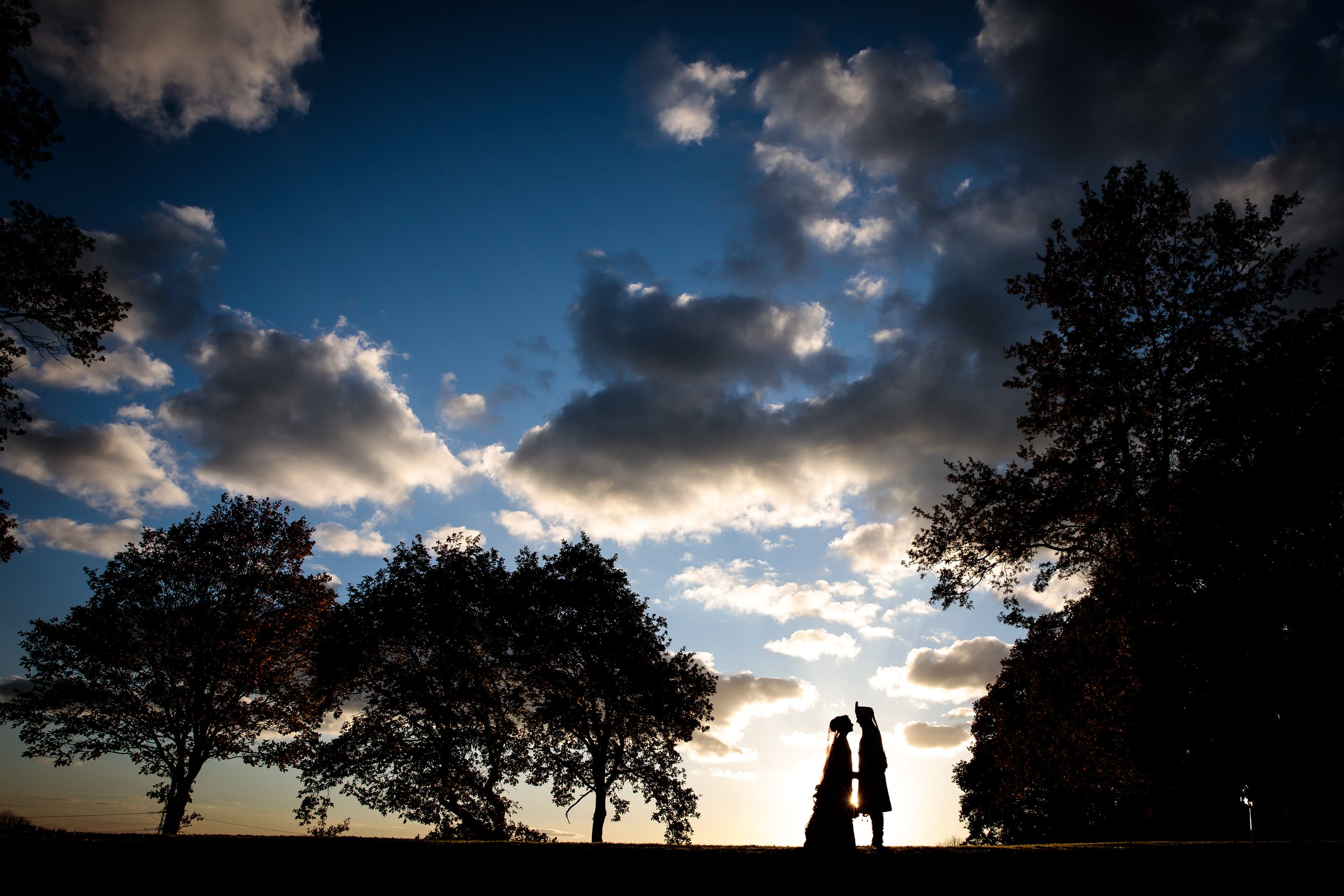 Silhouette of a couple standing on a hill with trees, looking at the sunset or sunrise sky filled with clouds.