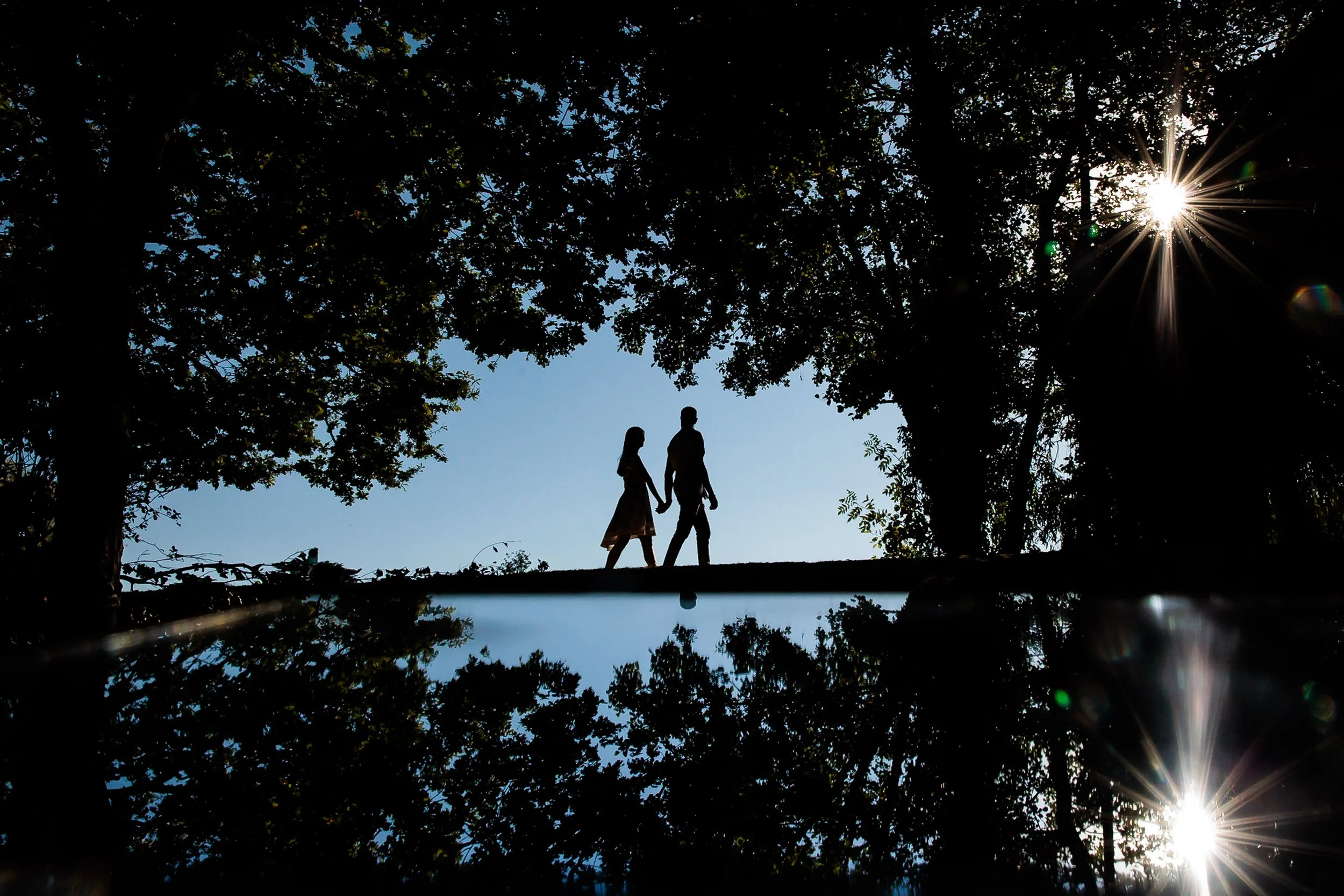Silhouettes of a man and woman holding hands, walking in a park with trees, with the sun shining through the branches, creating a starburst effect.
