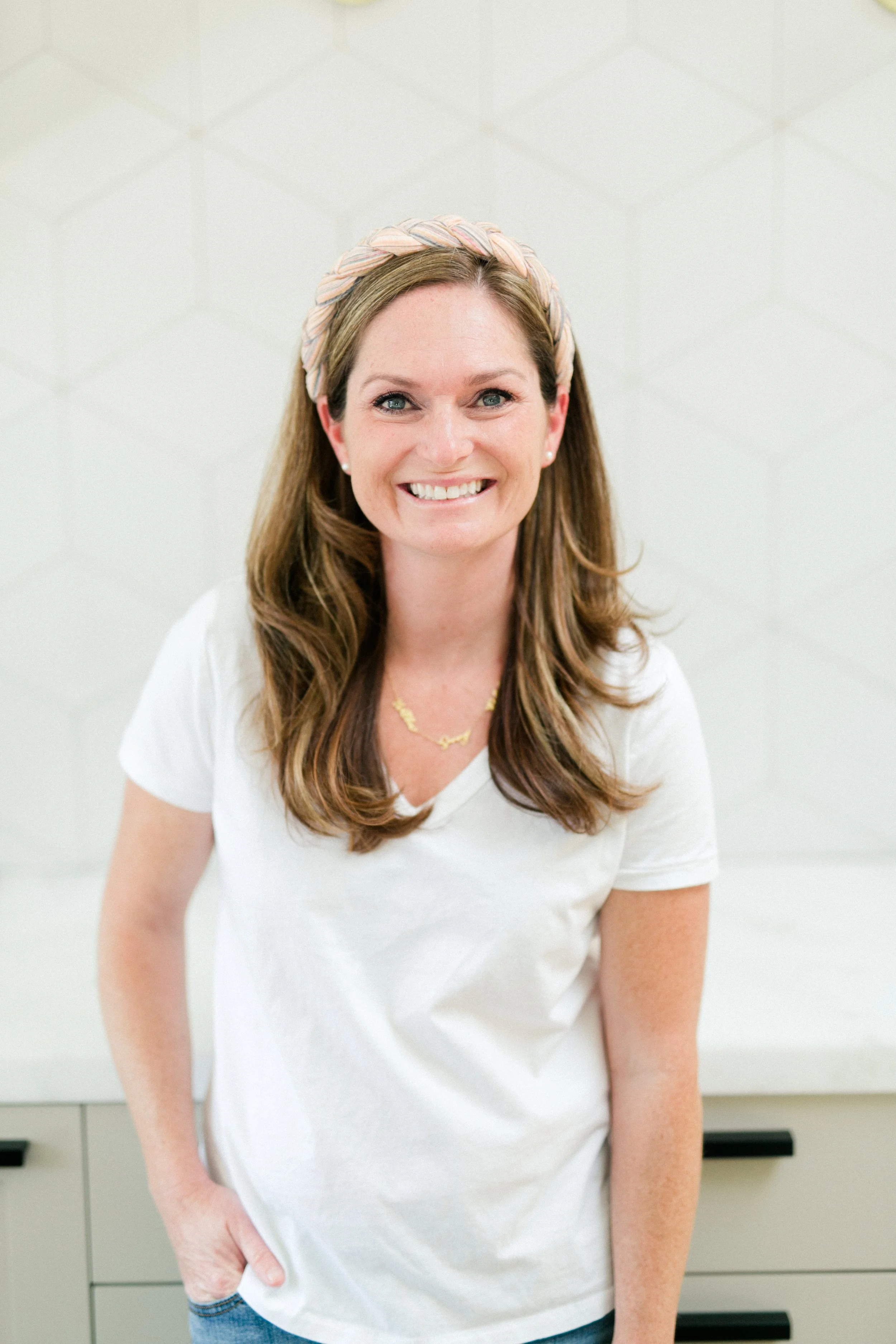 A smiling woman with brown hair wearing a white t-shirt, a light pink headband, and a gold necklace, standing against a white textured wall.