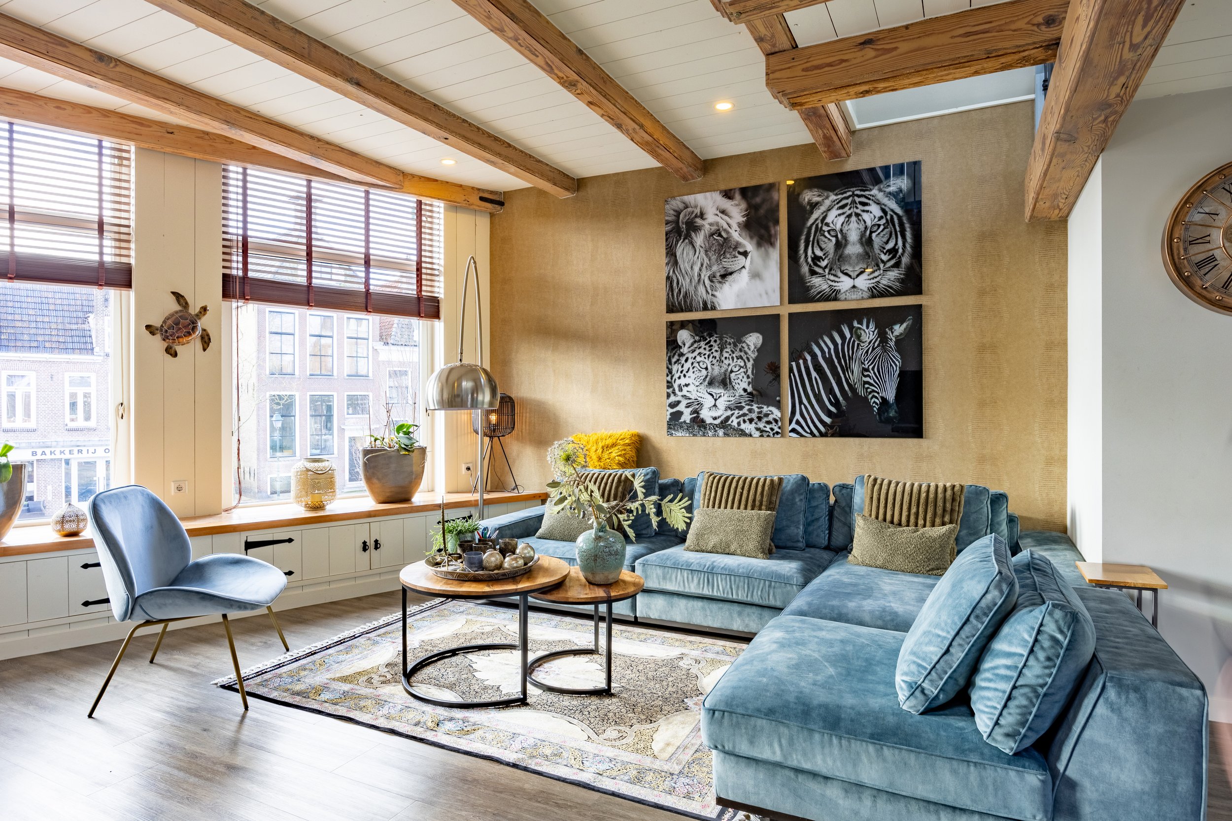 Een woonkamer met grote ramen, een blauwe bank en een enkele fauteuil, decoraties en planten op de vensterbank, en een muur met vier zwart-wit foto's van dieren. Er is een vloerkleed en een salontafel met decoratieve items.