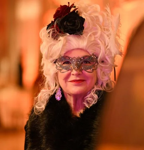 An elderly woman dressed in an elaborate costume with a curly white wig decorated with feathers and flowers, wearing a glittery mask and earrings, smiling at a masquerade party - Ballo dalle Rose.