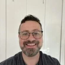 A smiling man with glasses, dark hair, and a beard, wearing a dark shirt, standing in front of a white wall.