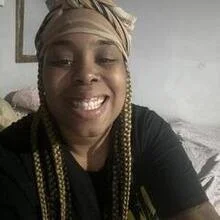 A woman smiling and wearing a headscarf or headwrap, with braided hair, indoors.