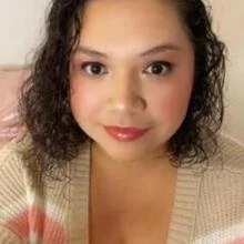 Close-up of a woman with curly dark hair and light makeup, wearing a beige sweater with pink and orange stripes, smiling softly at the camera.