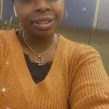Close-up of a smiling person wearing an orange sweater and a necklace with a heart pendant.