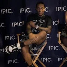 A man sitting on a chair with crossed arms and legs, wearing a black T-shirt, plaid shorts, and sneakers, in front of a backdrop with 'IPIC' logos.