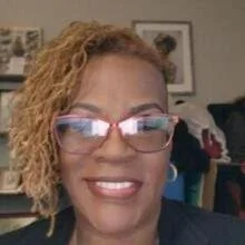 Close-up of a woman with curly blonde hair, glasses, and a big smile, in an indoor setting.