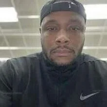A man wearing a black sports cap and black shirt taking a selfie indoors under fluorescent lights.