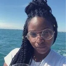 A woman with braided hair and glasses on a boat or near water, with a clear blue sky in the background.