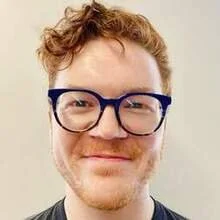Close-up of a young man with curly reddish hair and glasses, smiling slightly.
