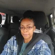 A woman with glasses and braided hair sitting in a car, wearing a plaid shirt and smiling slightly.
