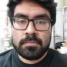 Close-up of a man with black hair, glasses, and a beard, looking directly at the camera.