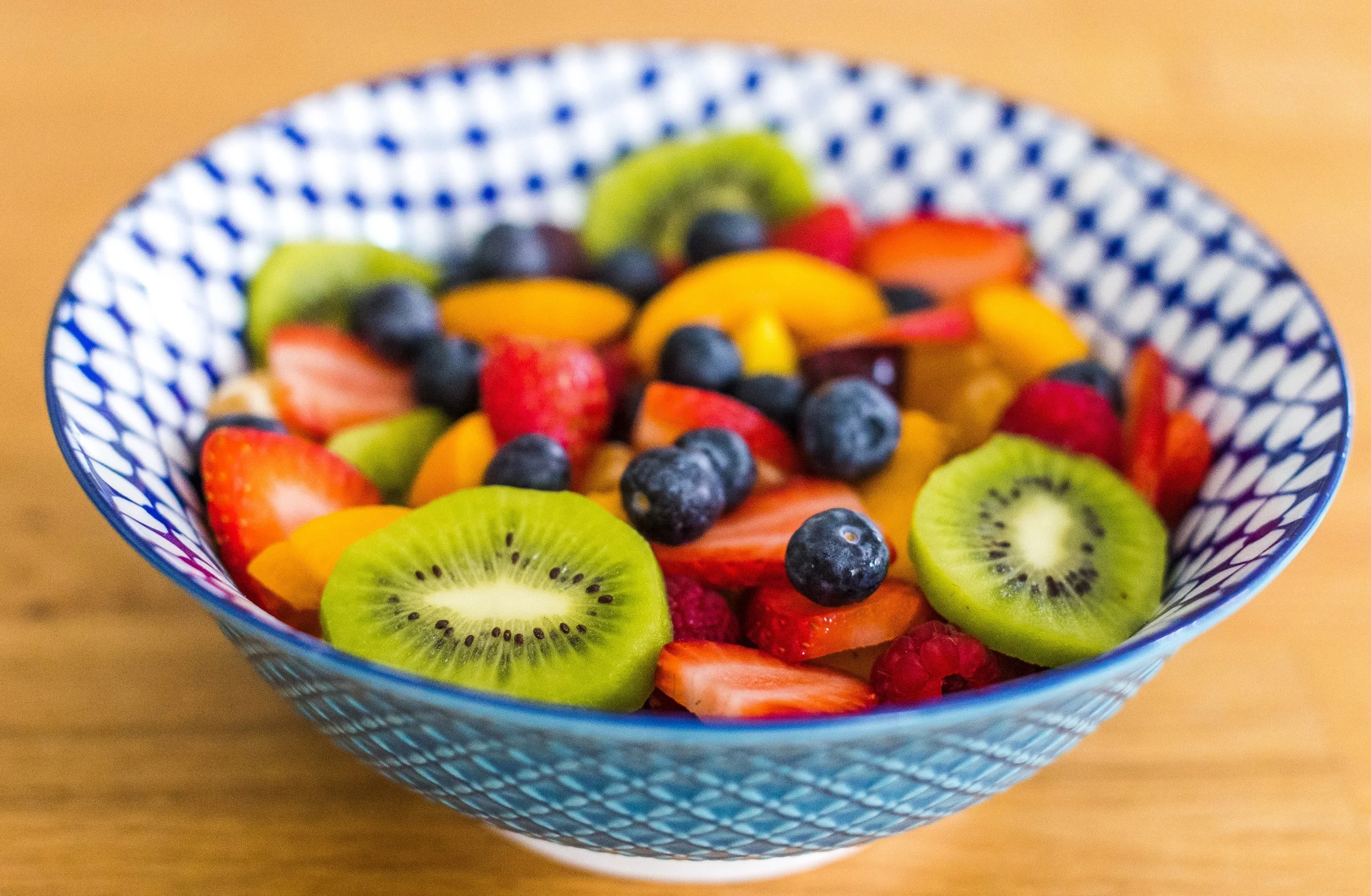 A bowl of mixed sliced fruits including kiwi, strawberries, blueberries, mango and peach slices in a blue patterned bowl.
